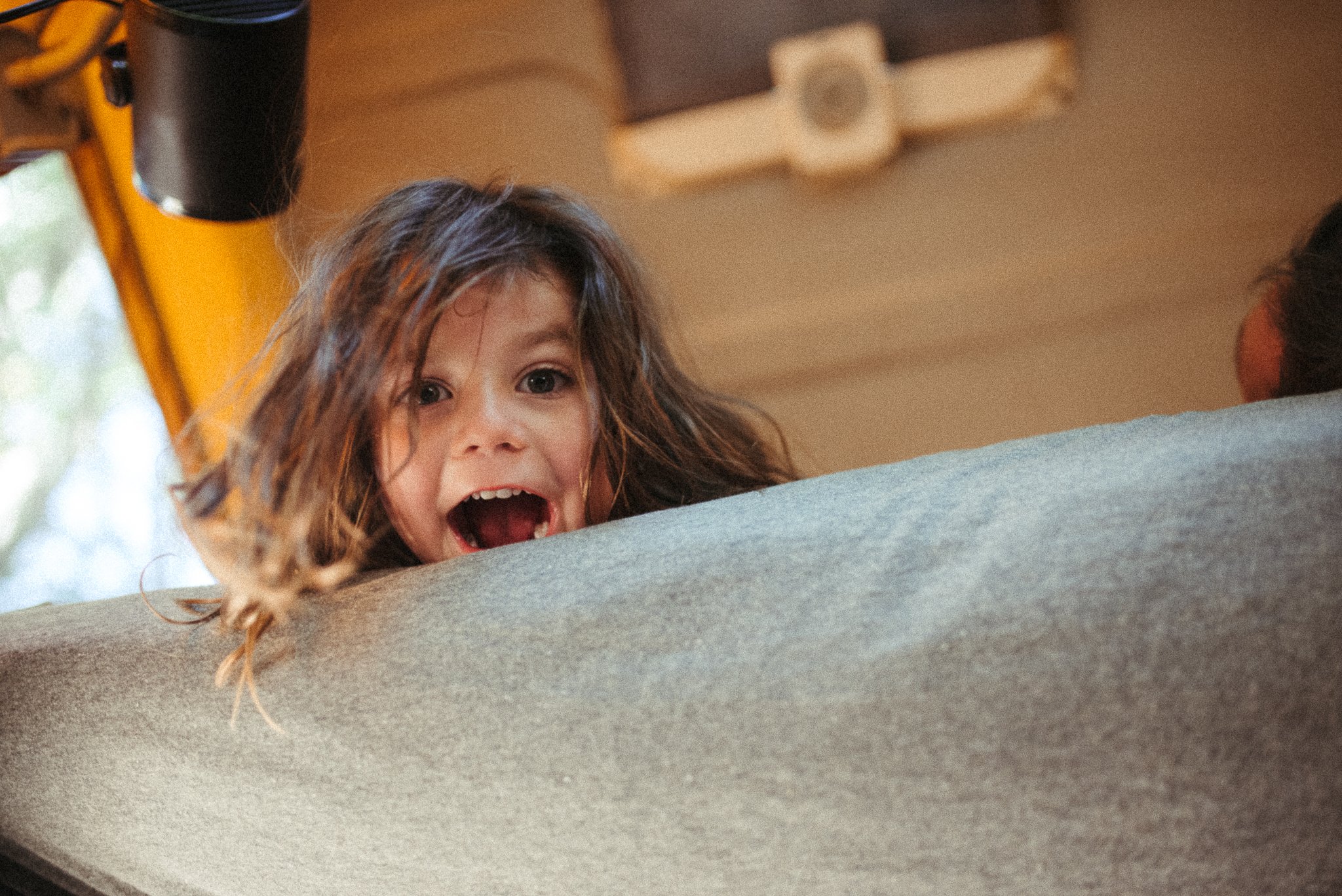A young girl with long, curly brown hair peeking over a gray surface, with a surprised or excited expression on her face