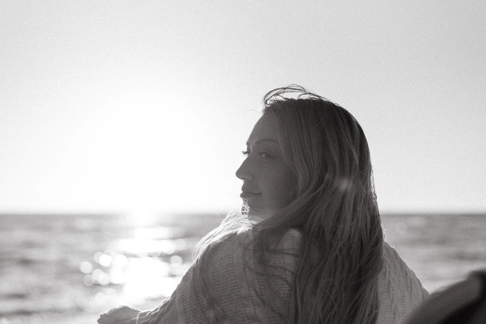 A woman with long hair, sitting and looking toward the ocean at sunset, in black and white.