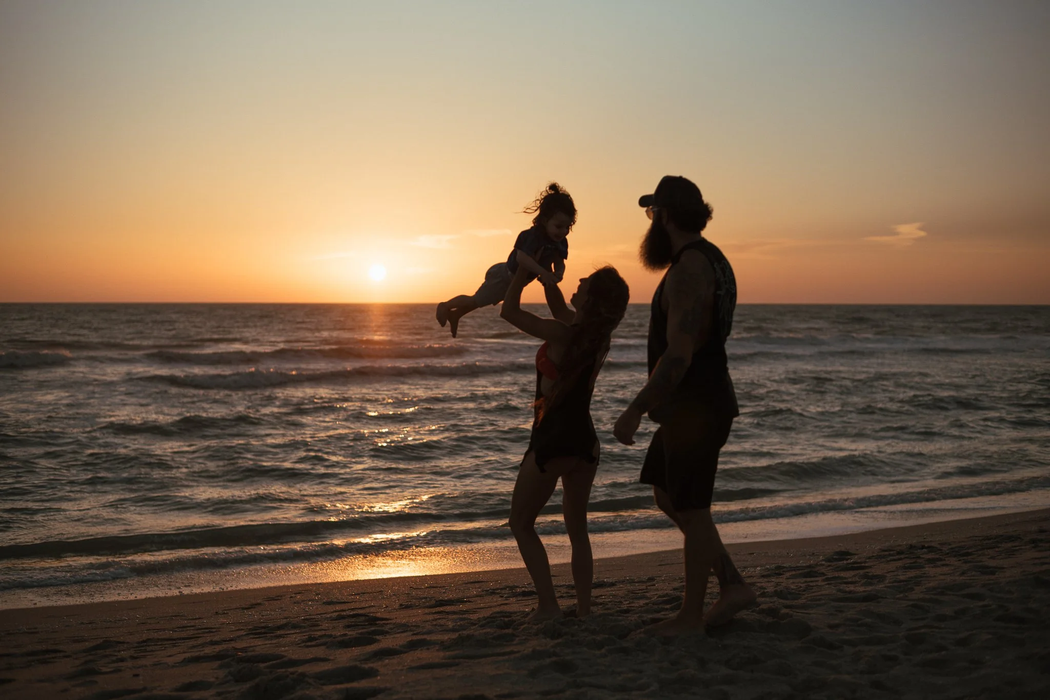 A family of three at the beach during sunset, with a man lifting a young girl in the air while the woman stands nearby, all silhouetted against the colorful sky and ocean.