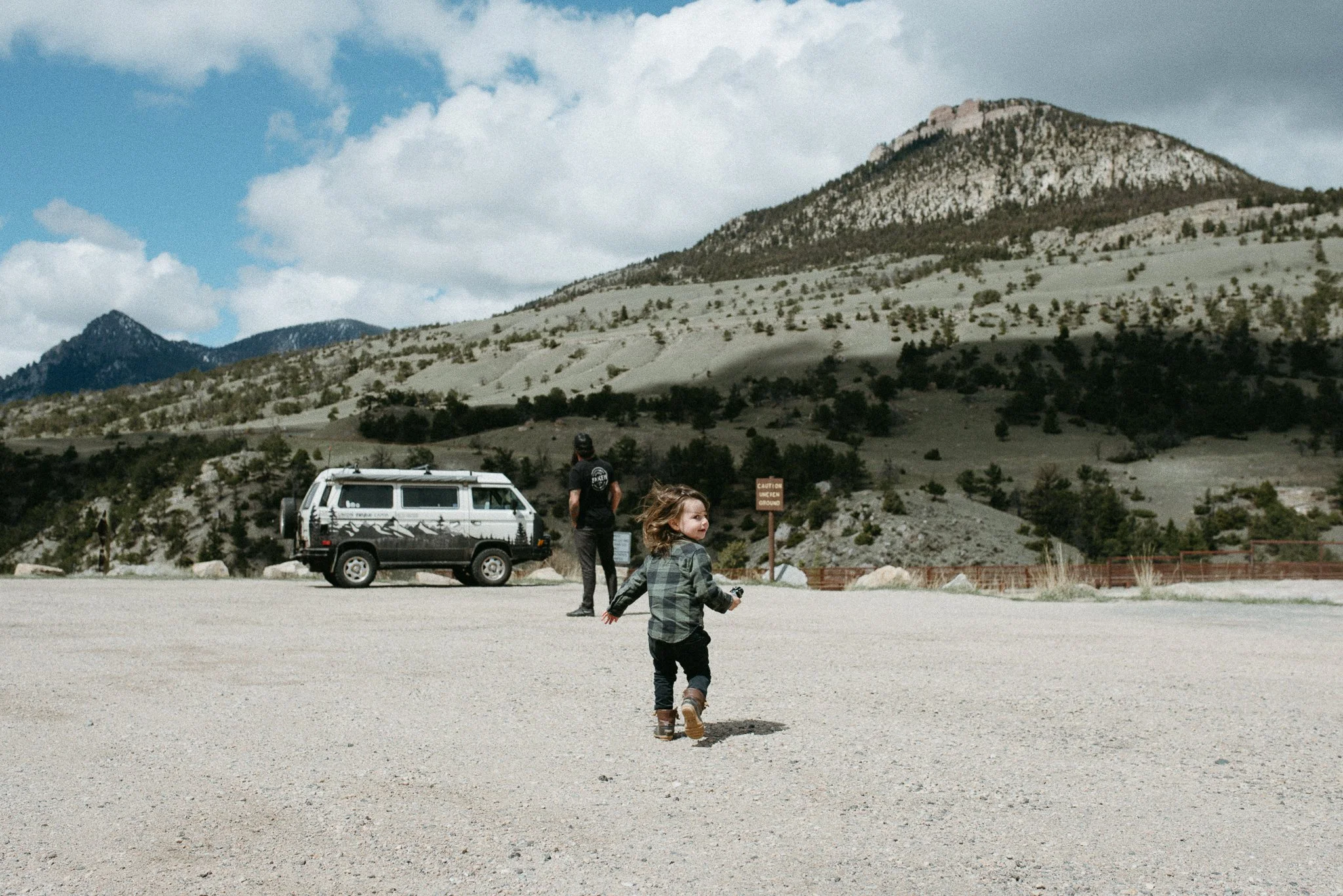 Two children playing in a wide open space with mountains and a partly cloudy sky in the background. A man stands near a vehicle, and there are signs and fencing in the distance.
