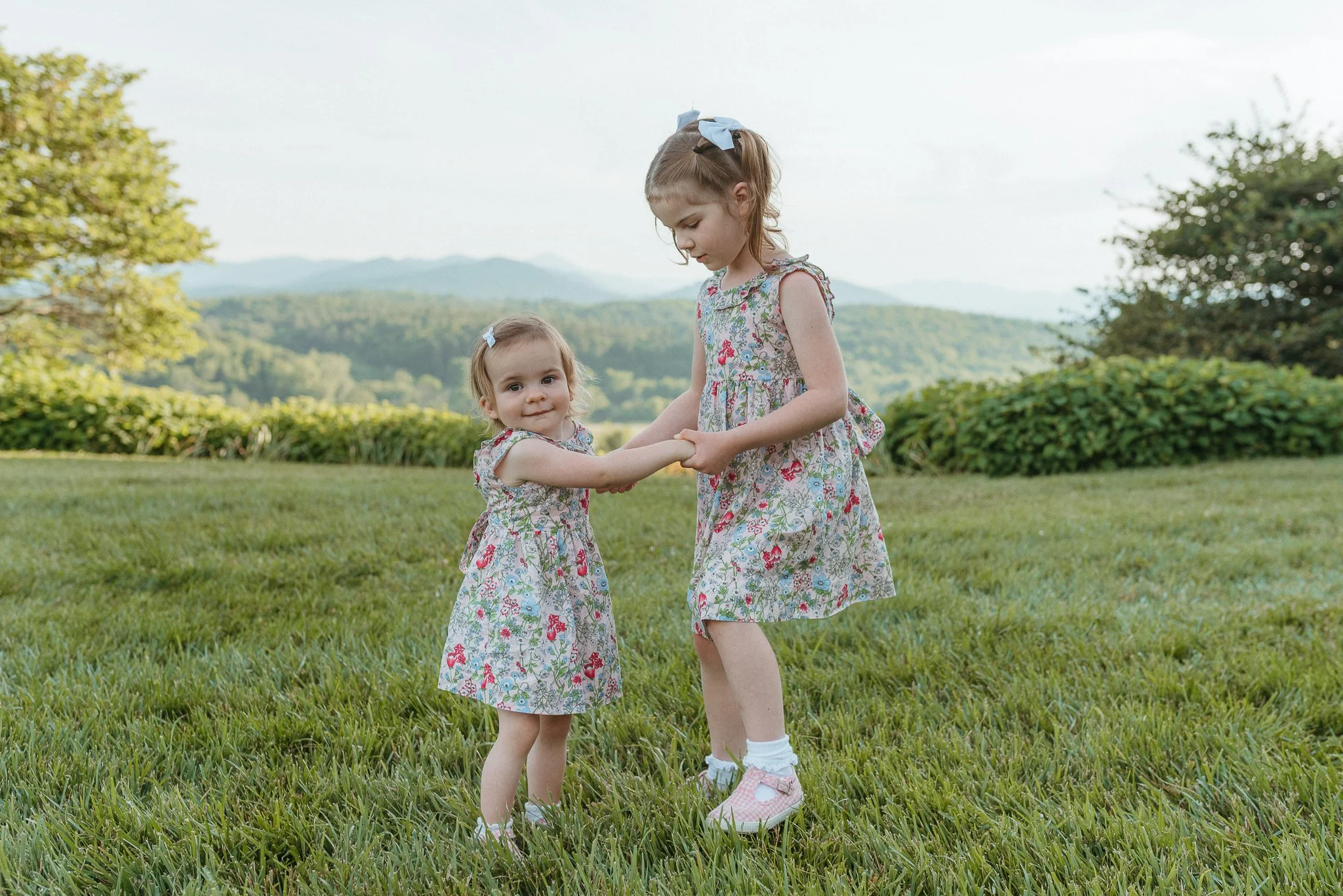 Two young girls in floral dresses holding hands and playing on a grassy field with mountains in the background.