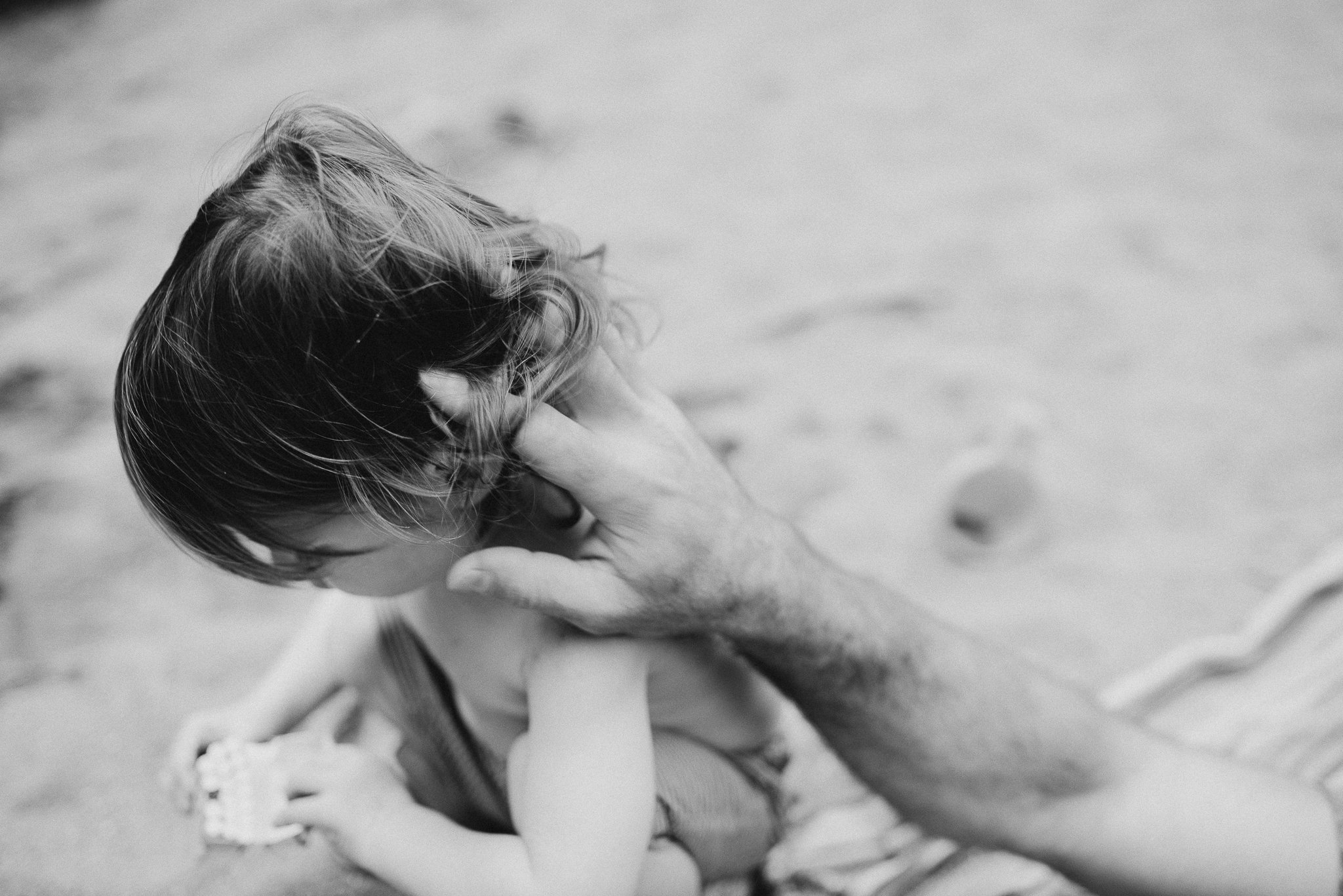 A person with long hair is giving a gentle kiss or nuzzle to a child with short hair on the beach, both facing each other.