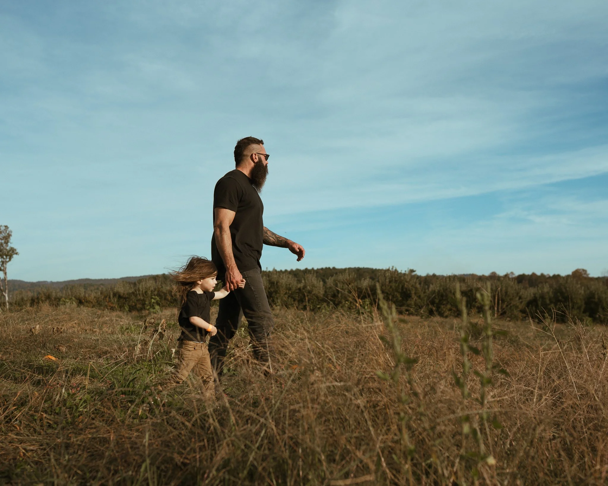 A man with a beard and glasses walking hand-in-hand with a young girl in a grassy, open field under a partly cloudy sky.