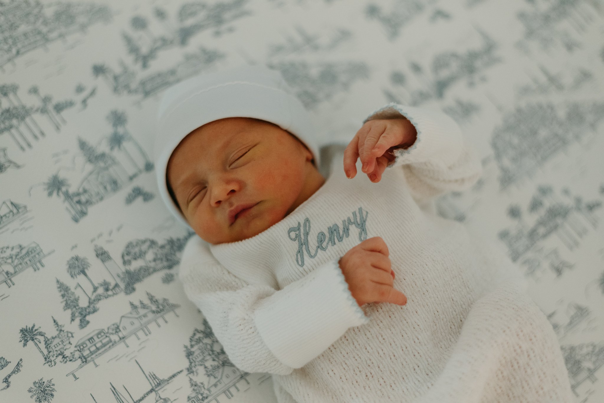 A sleeping newborn baby wearing a white hat and a white sweater with embroidery, lying on a blanket with a gray and white pattern of palm trees and buildings.