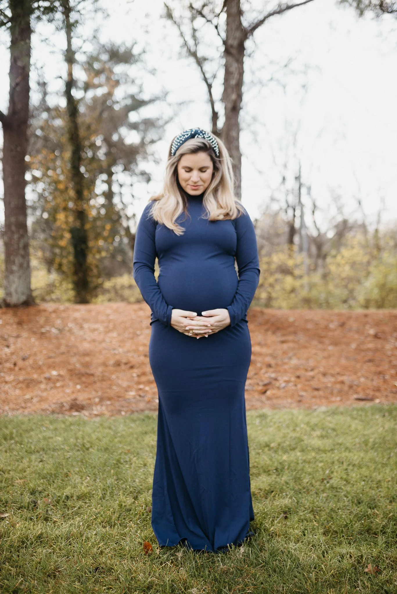 A pregnant woman in a navy blue dress standing outdoors with trees in the background, her eyes closed and her hands gently resting on her belly.