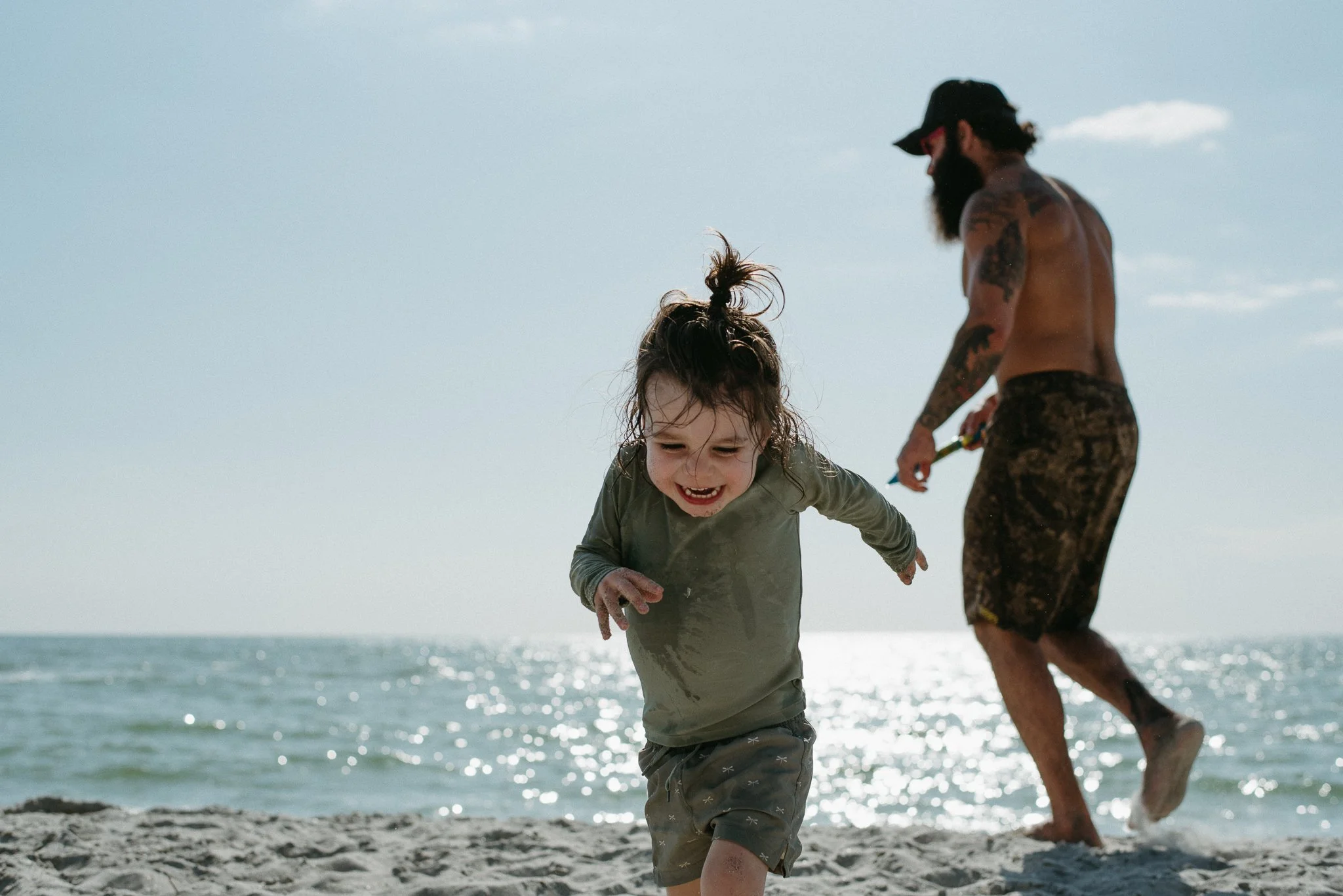 A young girl playing joyfully at the beach with an adult man, possibly her father, in the background. They are near the shoreline with water and sky visible, and both appear wet from playing in the water.