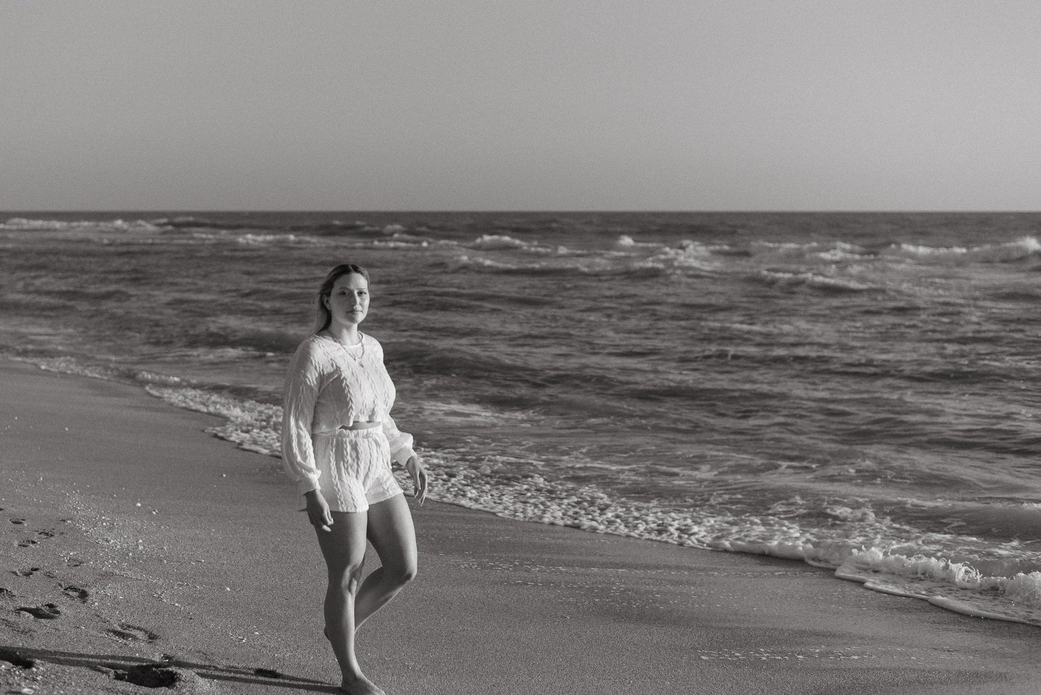 A woman in a light-colored dress standing on the sandy beach near the ocean, with waves in the background.