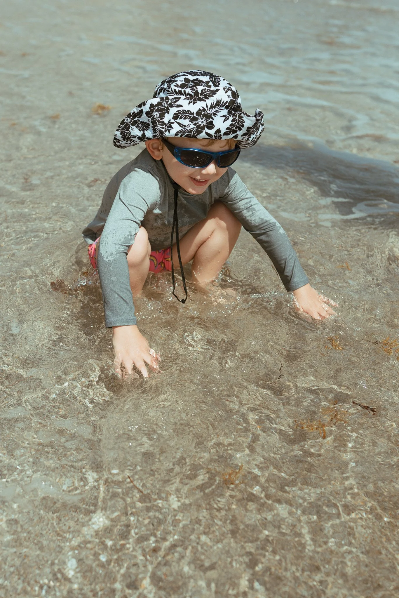 A young child wearing sunglasses, a sunhat, a long-sleeved shirt, and colorful shorts, crouching at the edge of a body of water, with hands in the water and smiling.