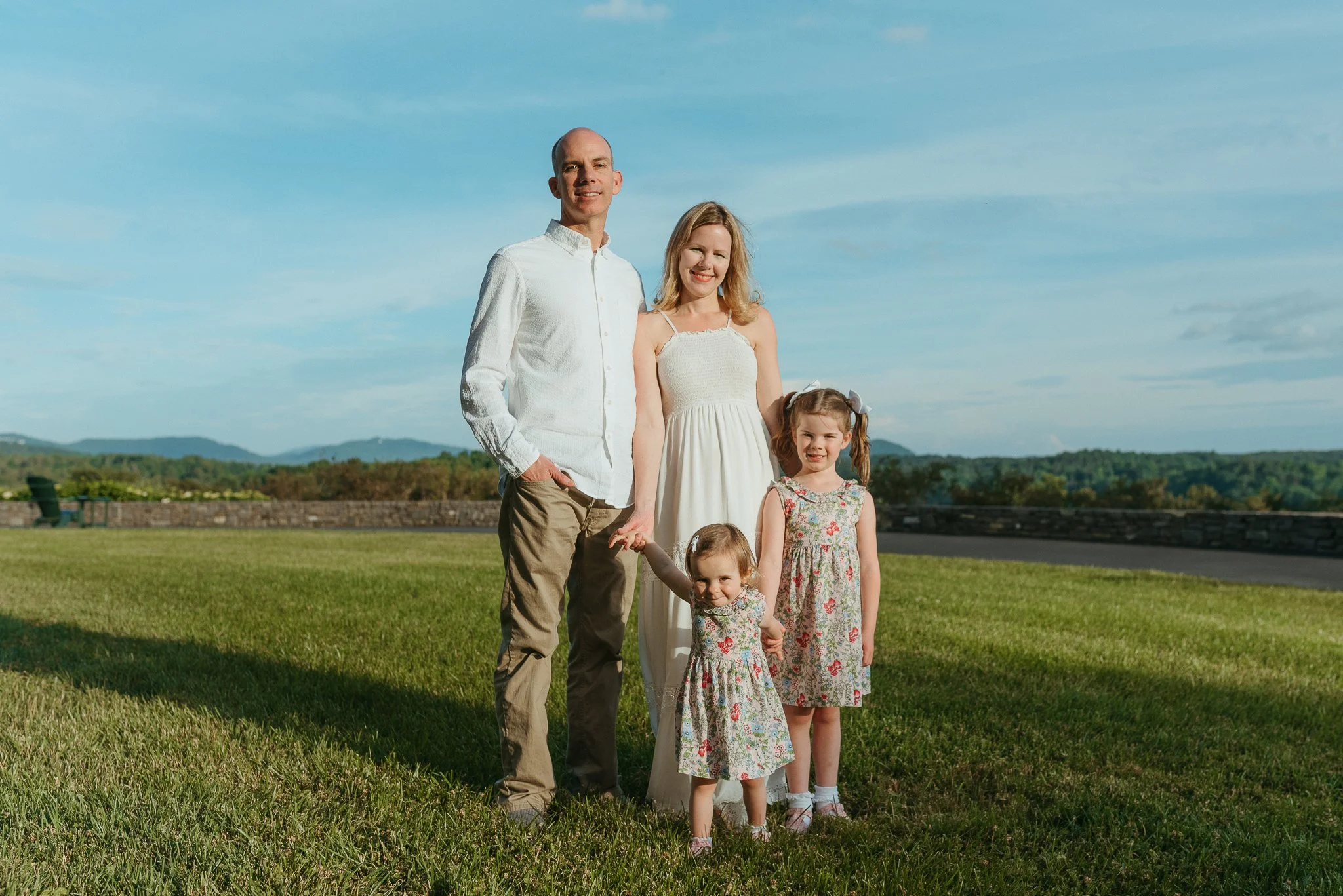 Family of six standing on a grassy park with mountains and blue sky in the background.