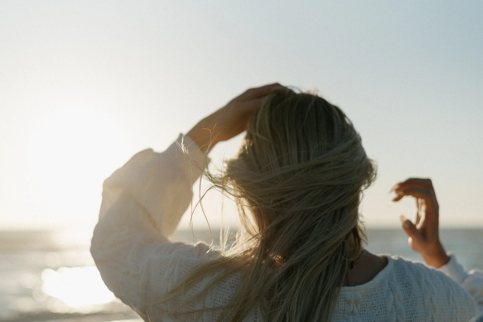 A person with long blond hair is outdoors, raising their hand to their head. The sky is clear and bright, suggesting a sunny day.