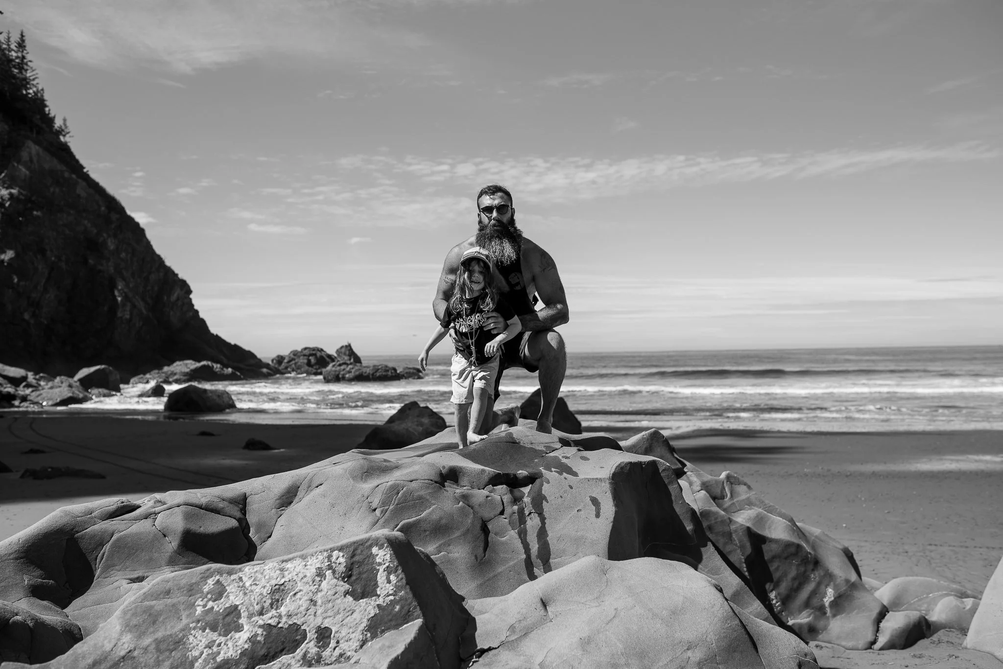 A man with a long beard, sunglasses, and tattoos kneels beside a young girl on a rocky beach.