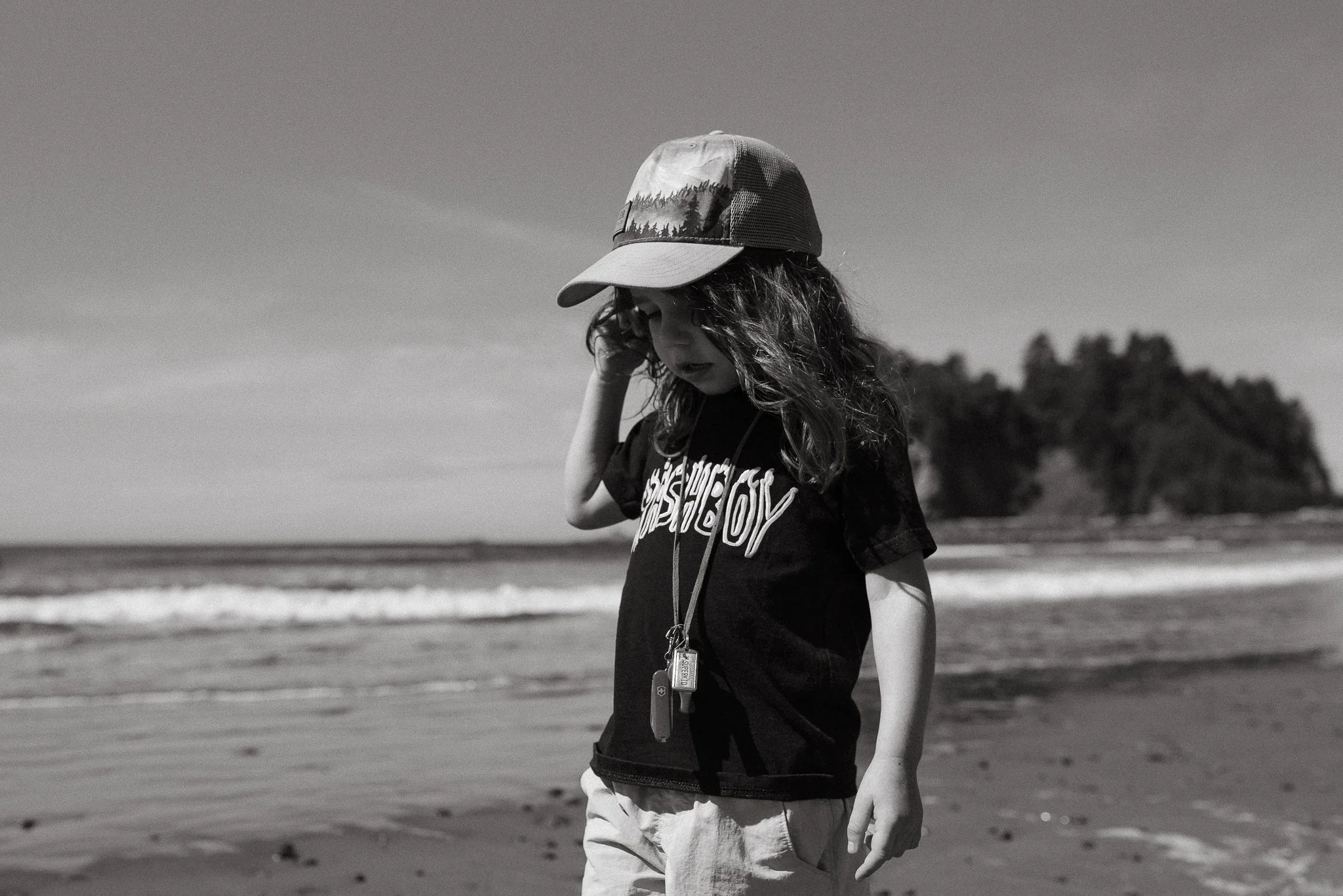 A young girl standing on the beach, wearing a cap, a graphic t-shirt, and shorts, with her head tilted downward and holding her head with one hand. The background features the shoreline and distant trees.