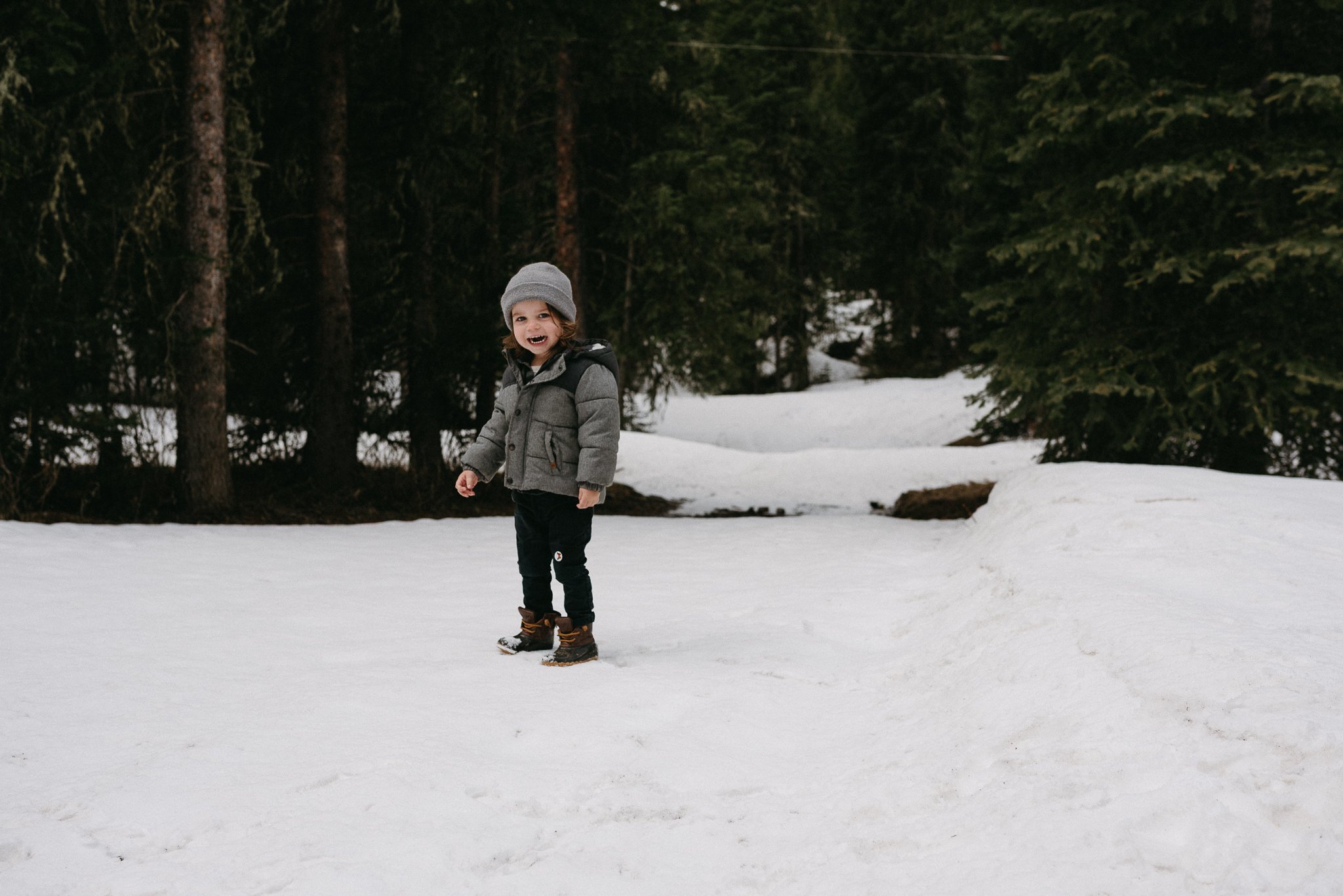 A young child with curly hair, wearing a gray beanie, gray jacket, black pants, and winter boots, standing on snow in a forested area with evergreen trees in the background.