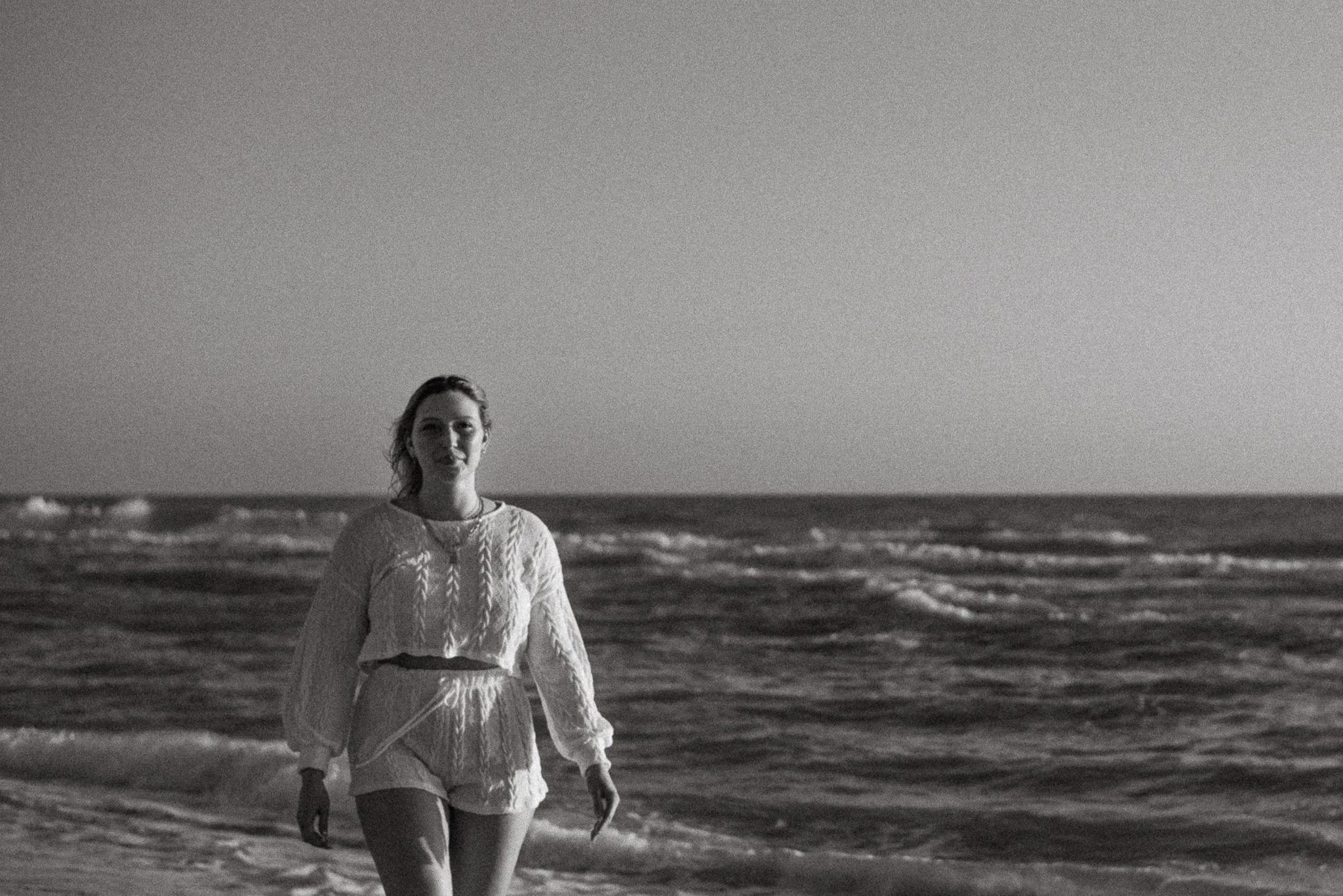 A woman is walking along the beach during what appears to be the evening or early morning, with the ocean and waves in the background. She is wearing a light-colored, textured sweater and light pants, and has hair that is wet or styled back.