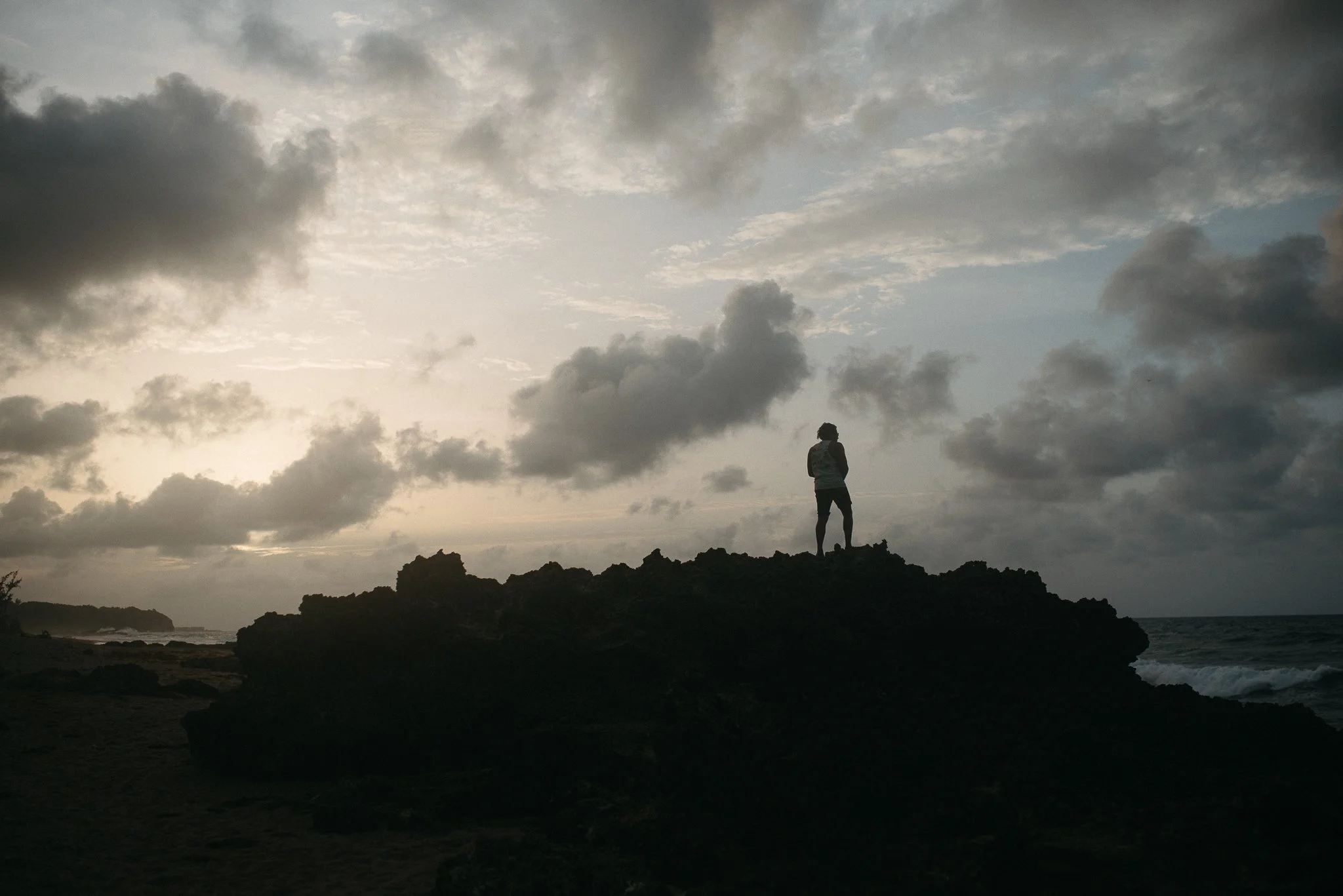 Person standing on rocky shoreline at sunset or sunrise, silhouetted against a cloudy sky.