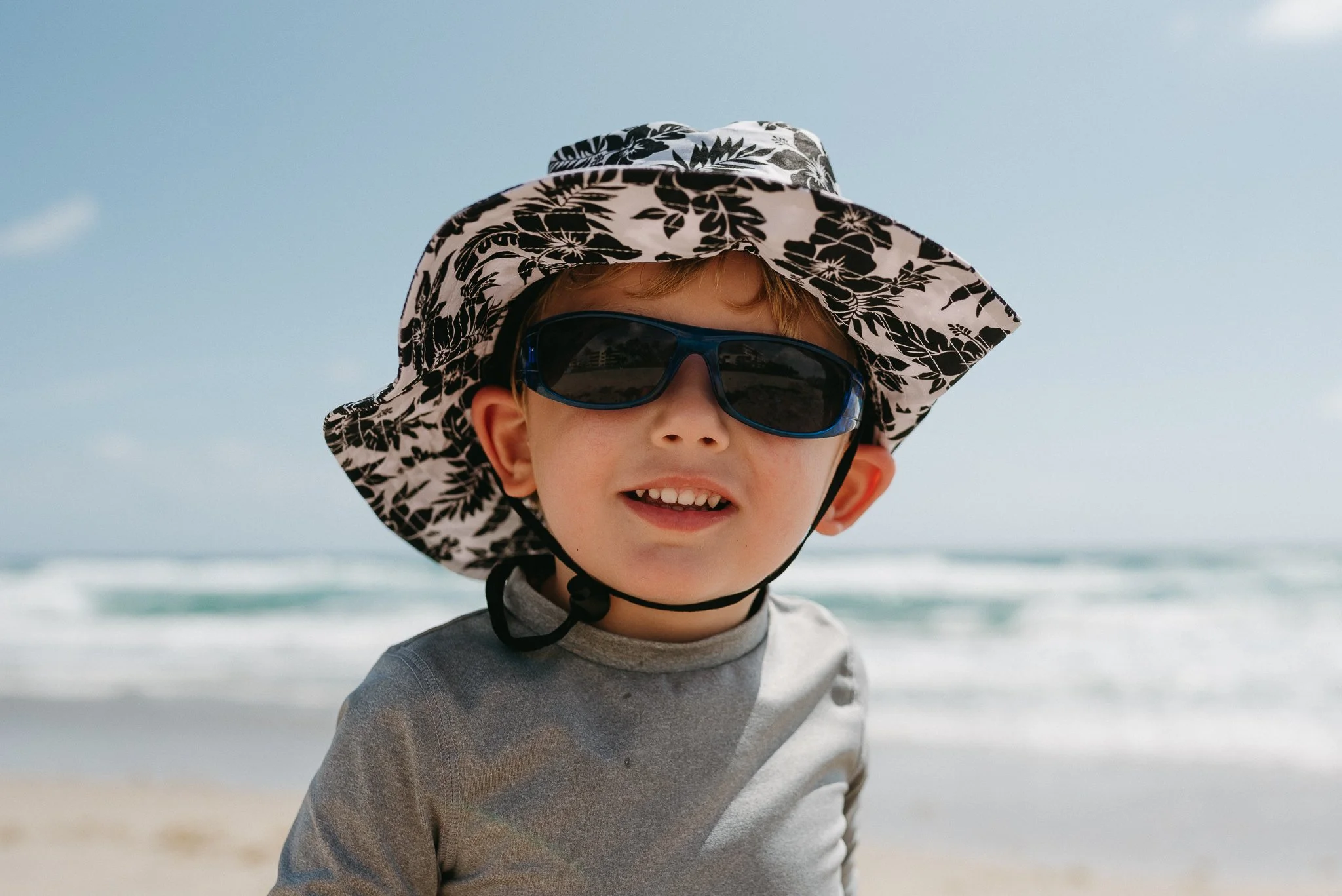 A young boy wearing a wide-brimmed hat with a black and white floral pattern and sunglasses at the beach.