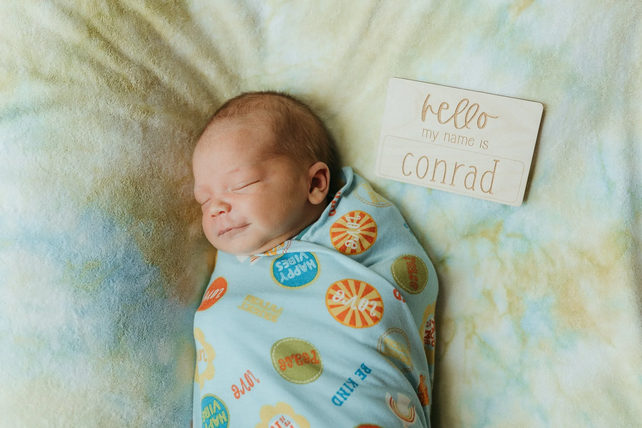 A sleeping baby wearing a colorful pajama with print of candies and the words 'Happy Vibes'. There is a small sign next to the baby that says 'Hello, my name is Conrad' on a soft, colorful blanket.