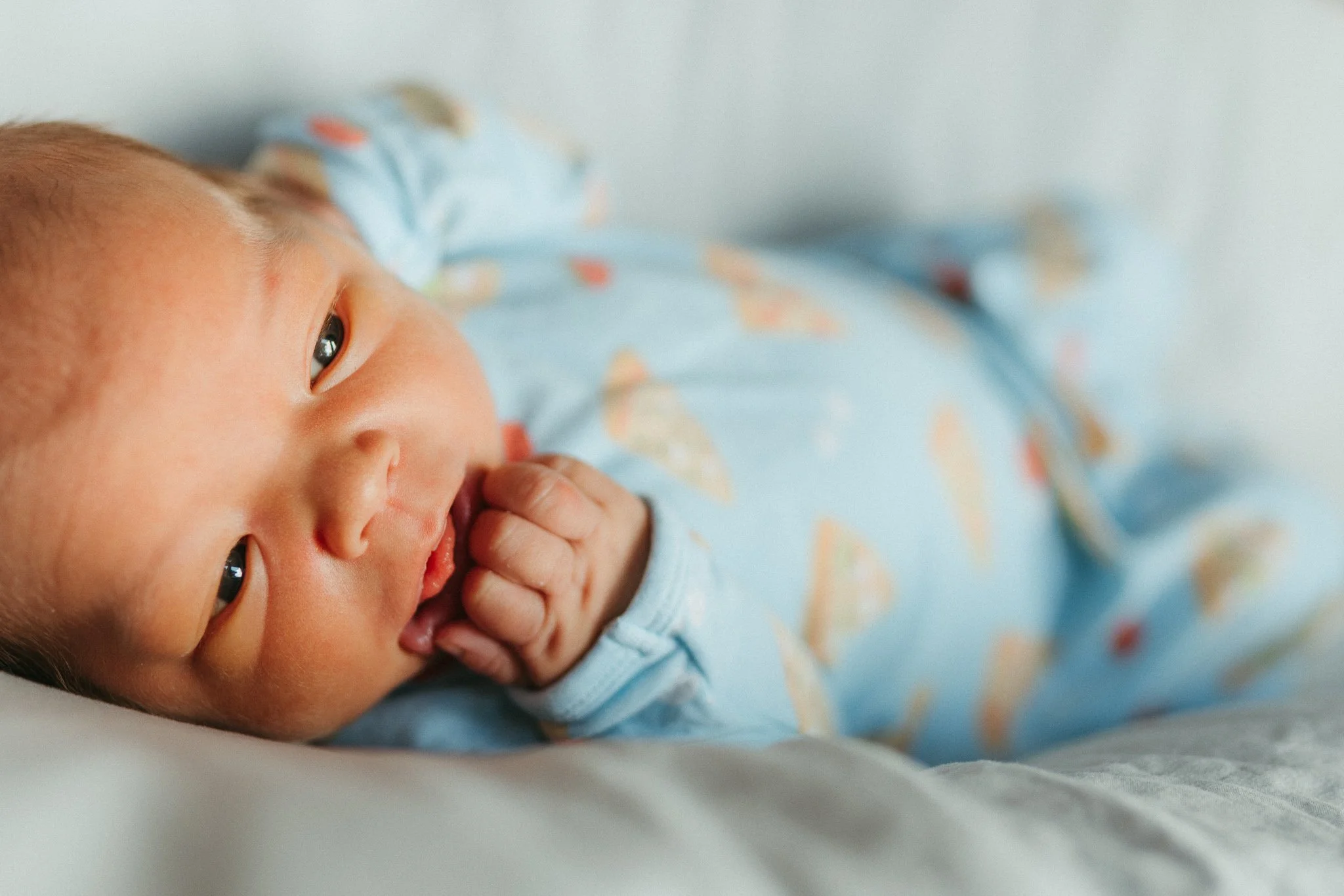 Close-up of a sleeping baby with eyes slightly open, lying on a bed or soft surface, wearing a light blue onesie with colorful patterns.