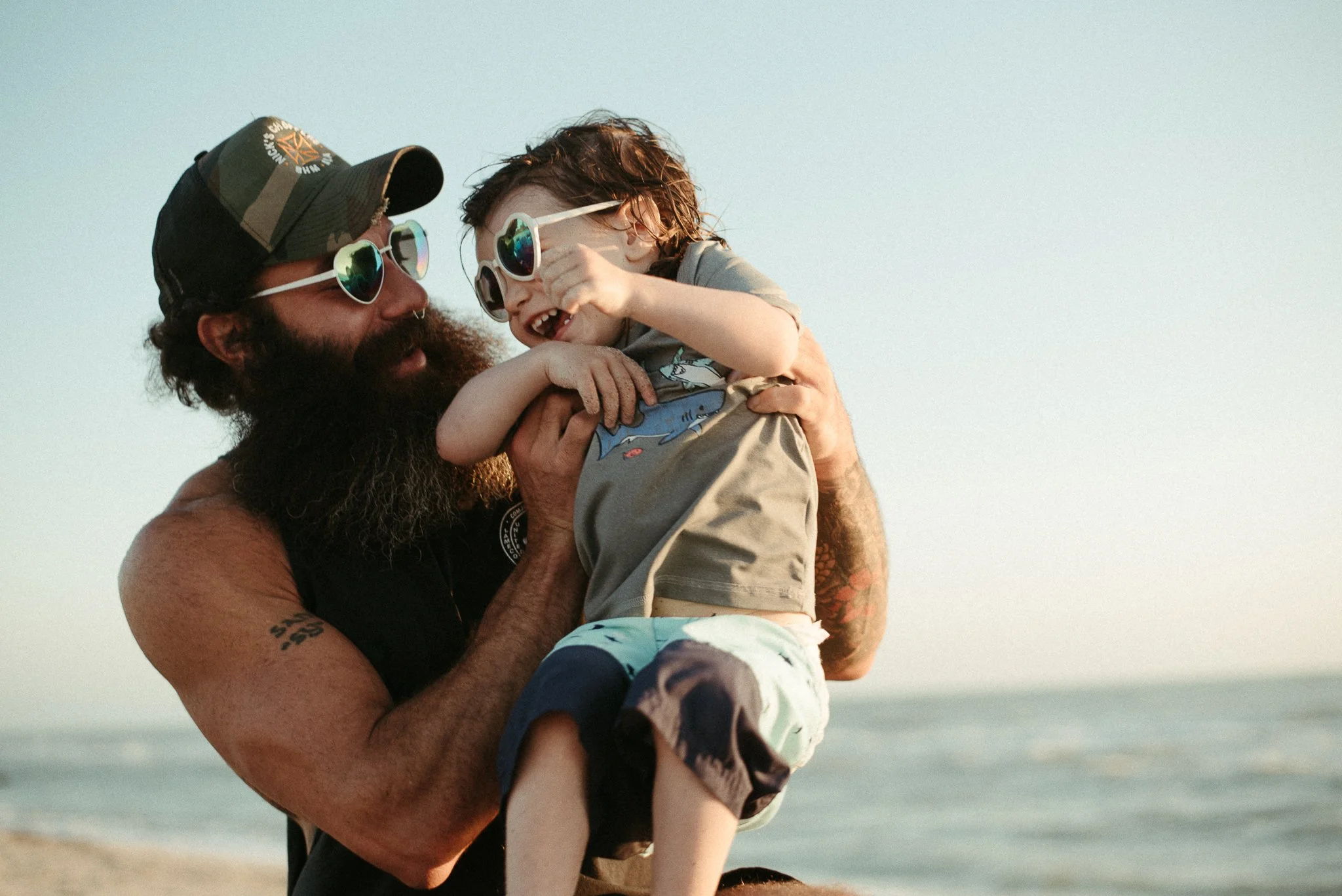 A man with a beard and sunglasses holding a young boy with sunglasses at the beach.