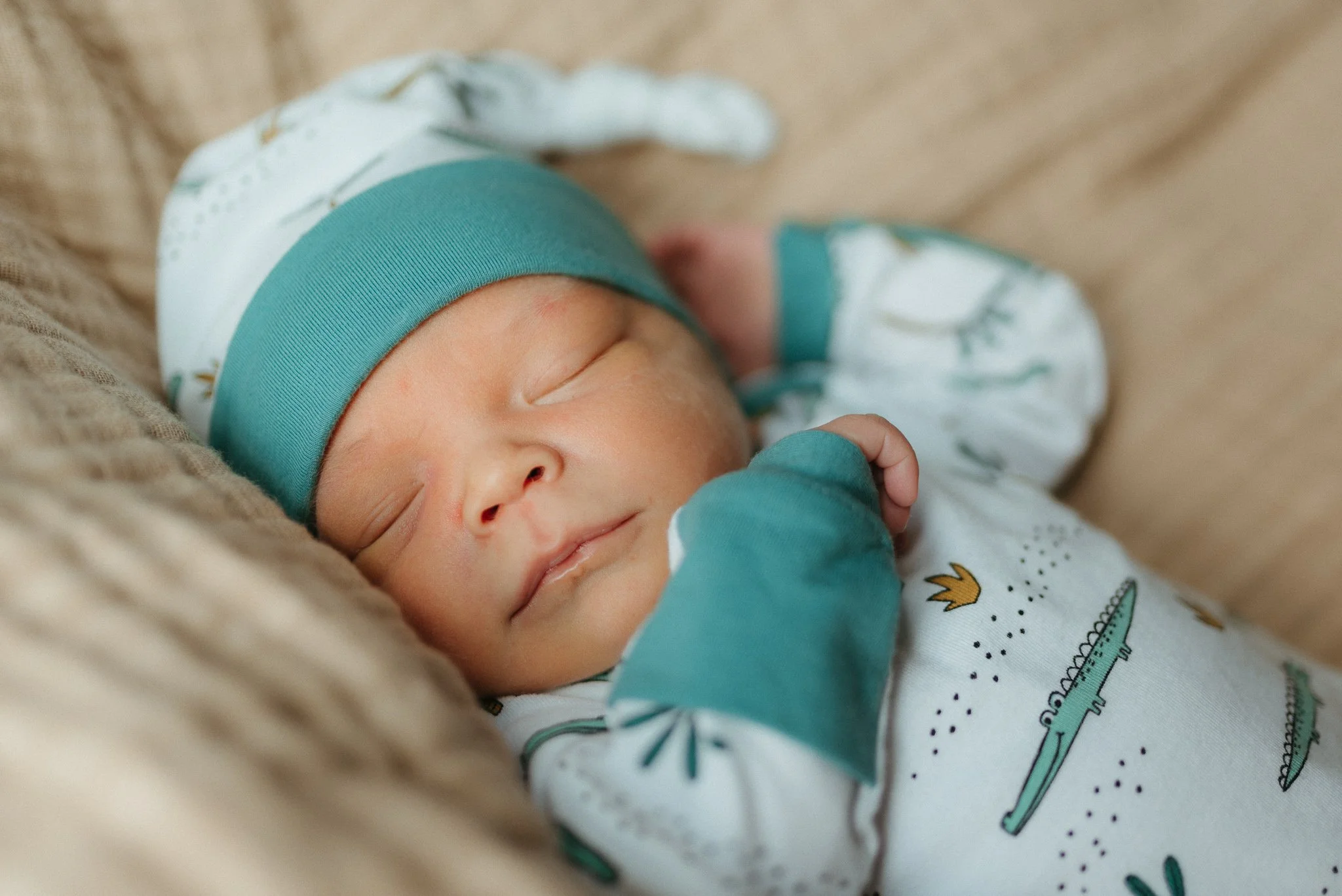 Close-up of a sleeping newborn baby wearing a teal and white hat and a printed onesie, lying on a soft surface.