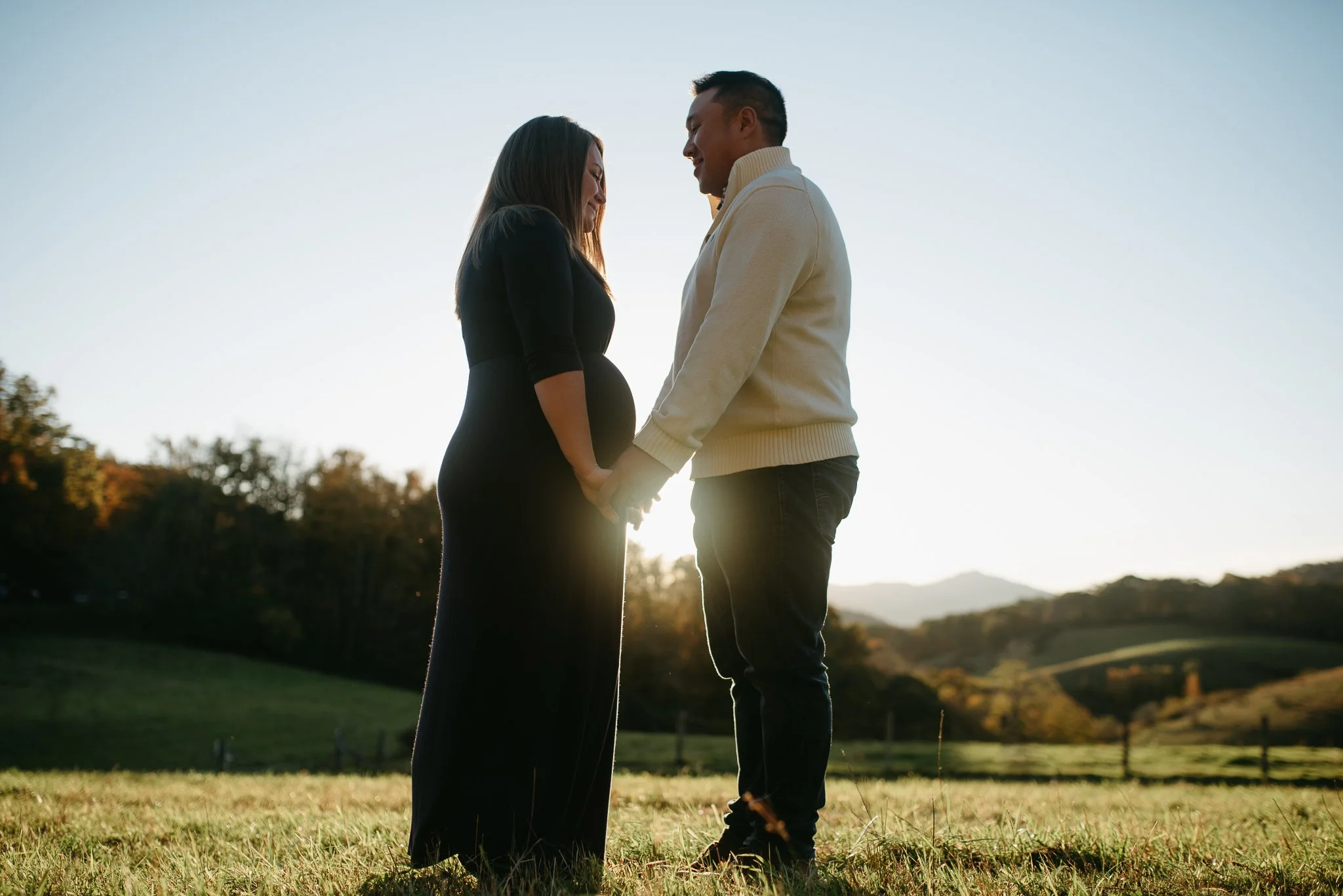 A pregnant woman and a man standing close together outdoors, holding hands and smiling at each other during sunset.