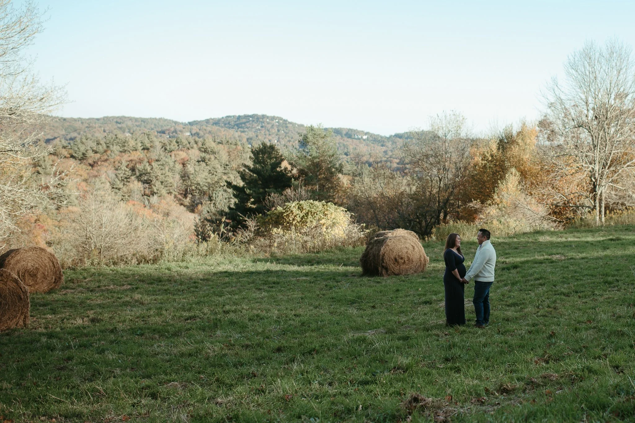 A couple standing in a grassy field holding hands, with hay bales and autumn trees in the background.