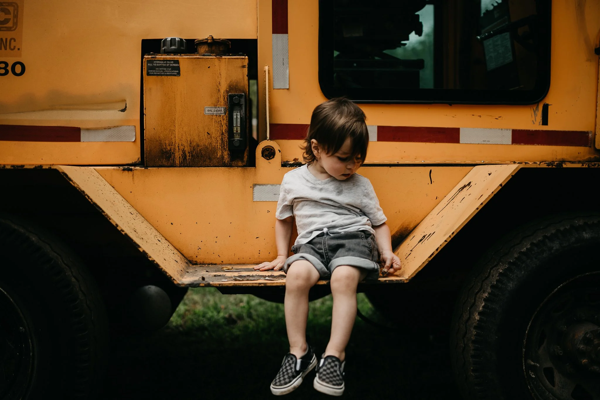 A young child with dark hair, wearing a light gray t-shirt, black shorts, and black-and-white checkered shoes, sitting on the step of a yellow construction vehicle or heavy machinery, with large black tires visible underneath.