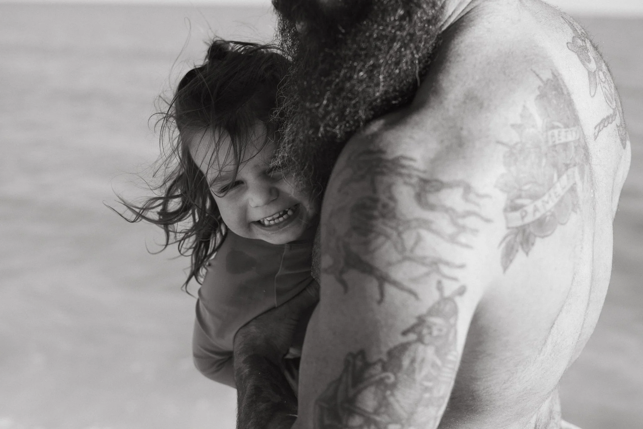 A young girl with curly hair smiling and hugging a person with tattoos, on a textured surface, shown in black and white.