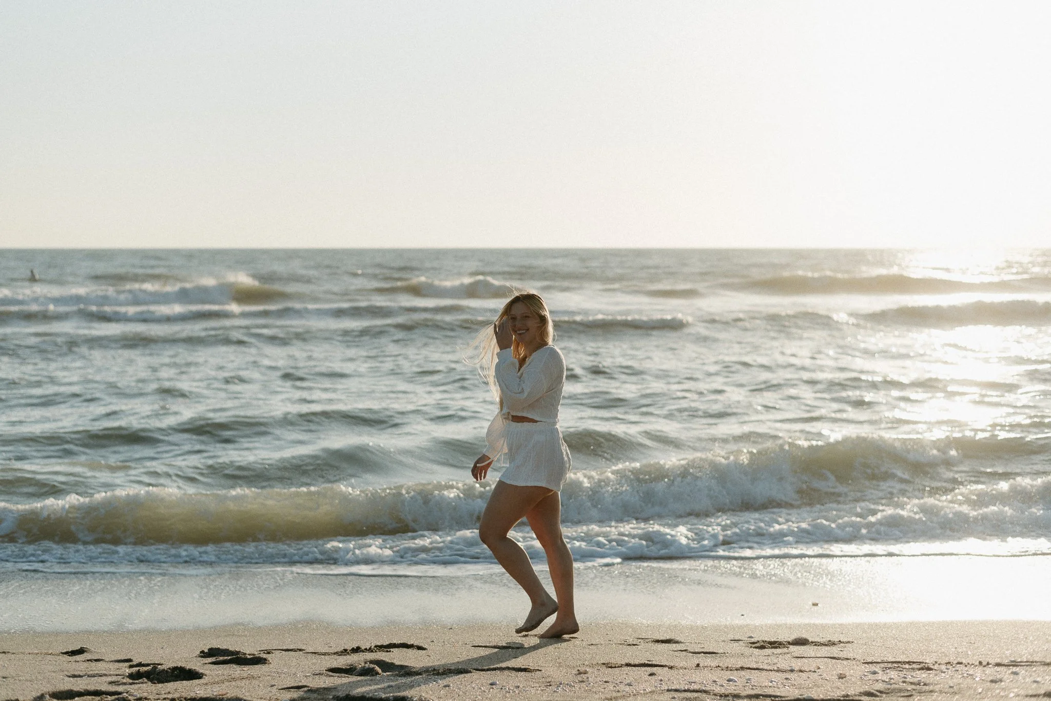 A woman in a white dress smiling and walking along the shoreline at the beach during sunset.