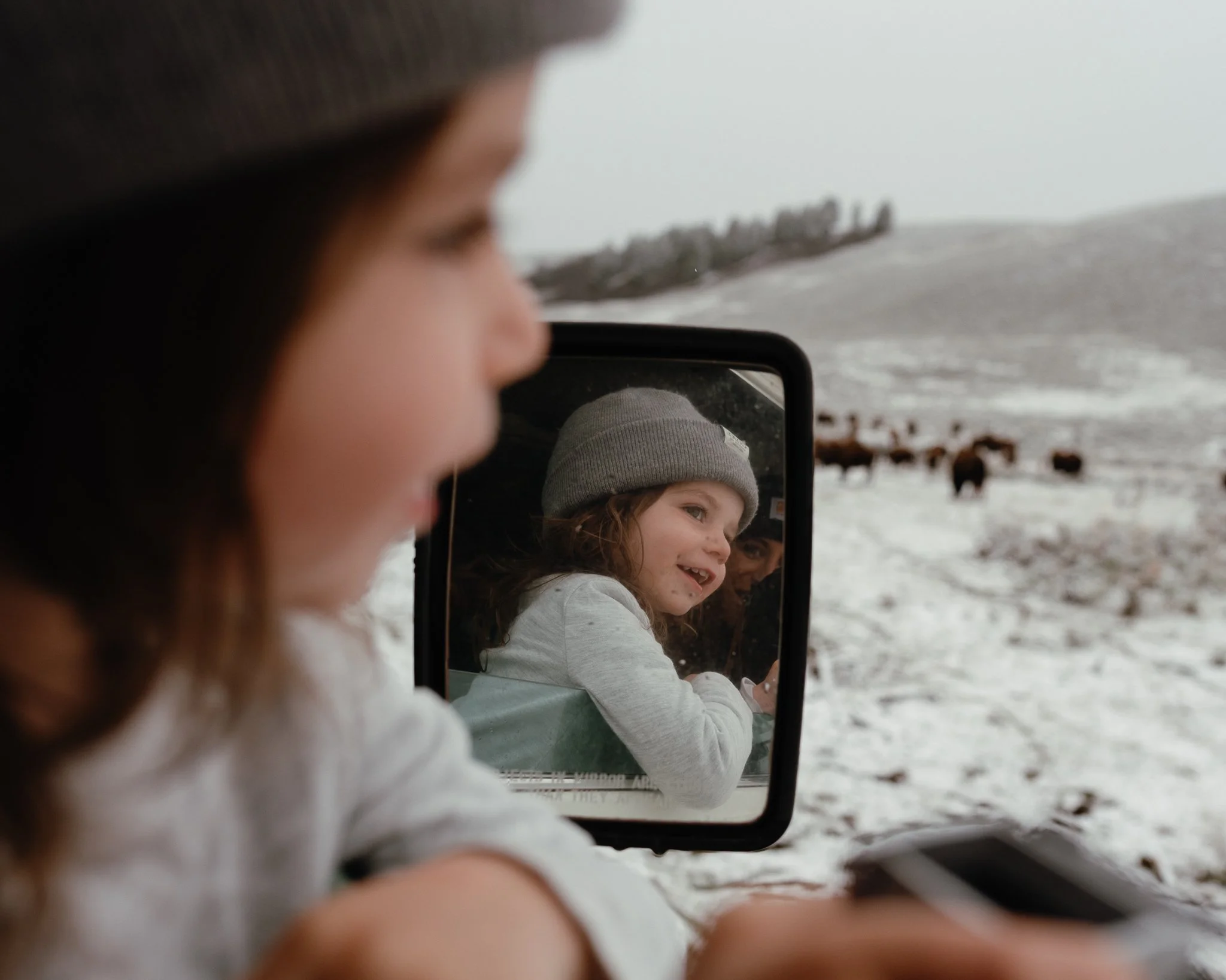 A young girl in a gray beanie is reflected in a mirror held by a stranger, with snow-covered landscape and cows in the background.