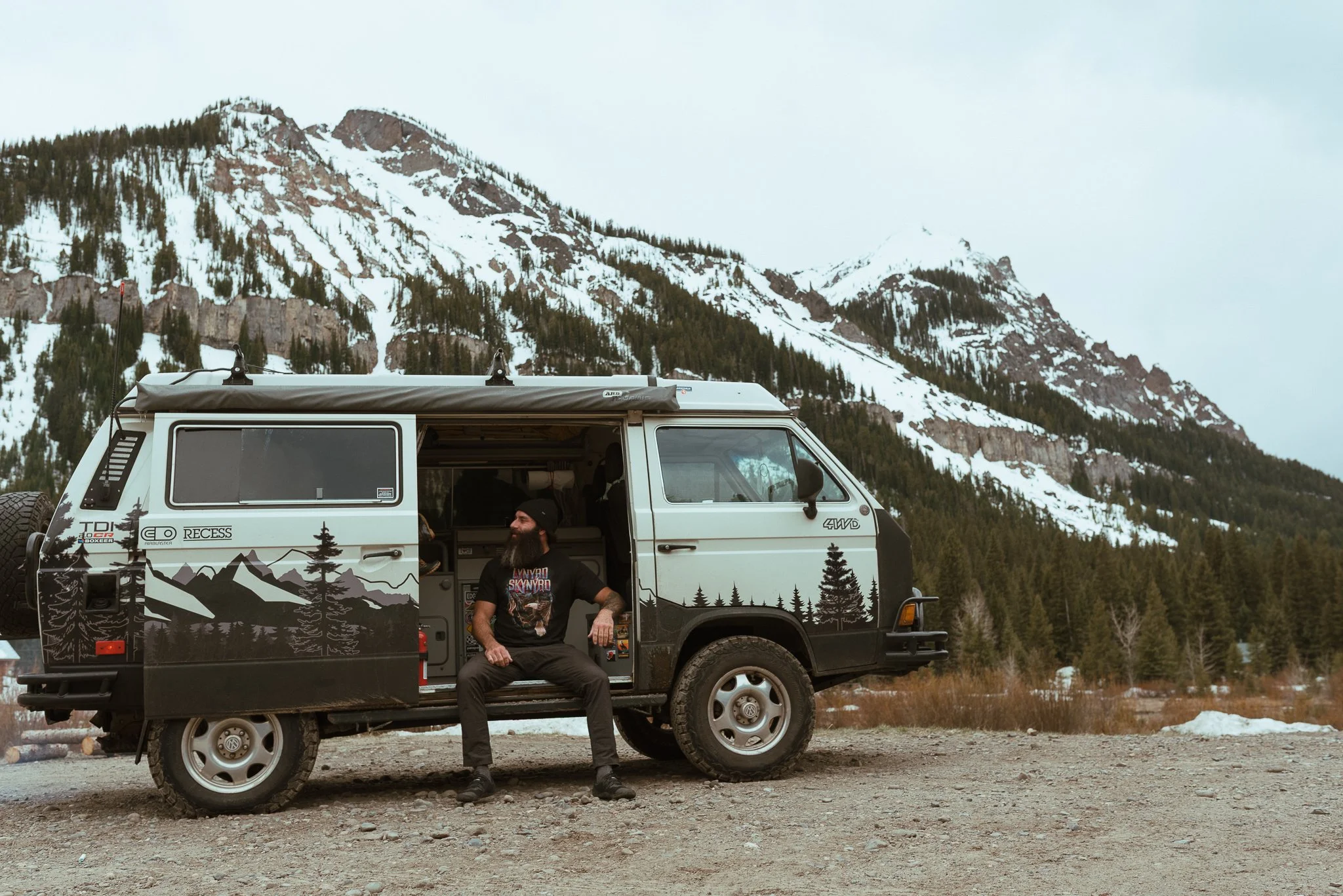 A man with a beard wearing a graphic t-shirt sitting on the open door of a camper van decorated with mountain and forest illustrations, parked on a gravel area with snow patches, and snow-capped mountains with pine trees in the background.