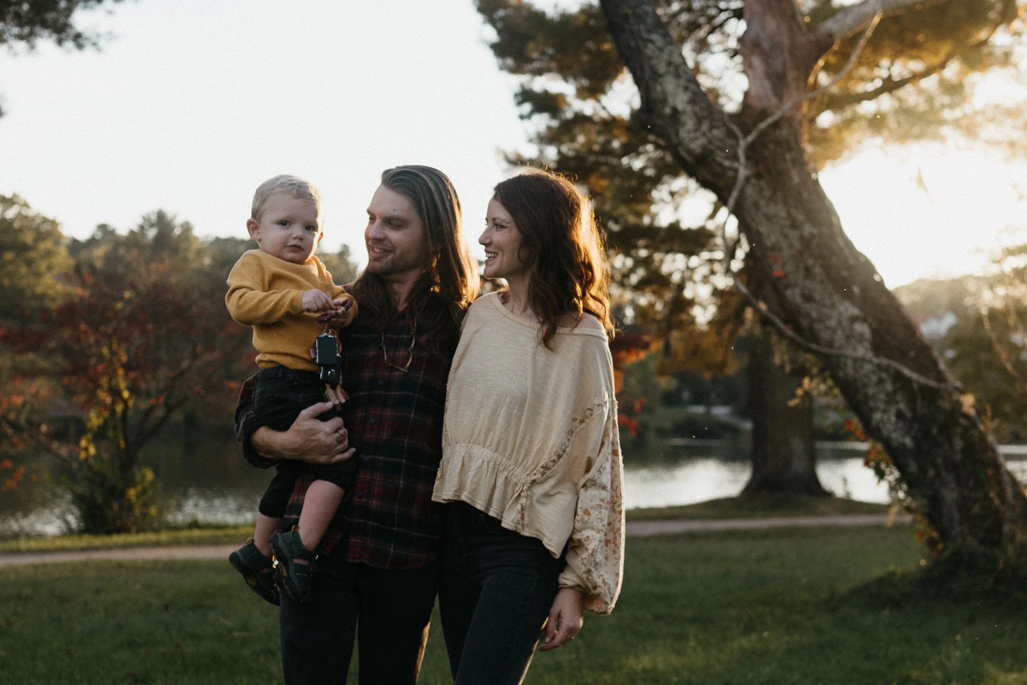 A family of three stands outdoors near a large tree during sunset. The man, with long hair, is holding a young child in a mustard sweater, and a woman with wavy hair is smiling at them. The background features a park with a pond and trees with autumn