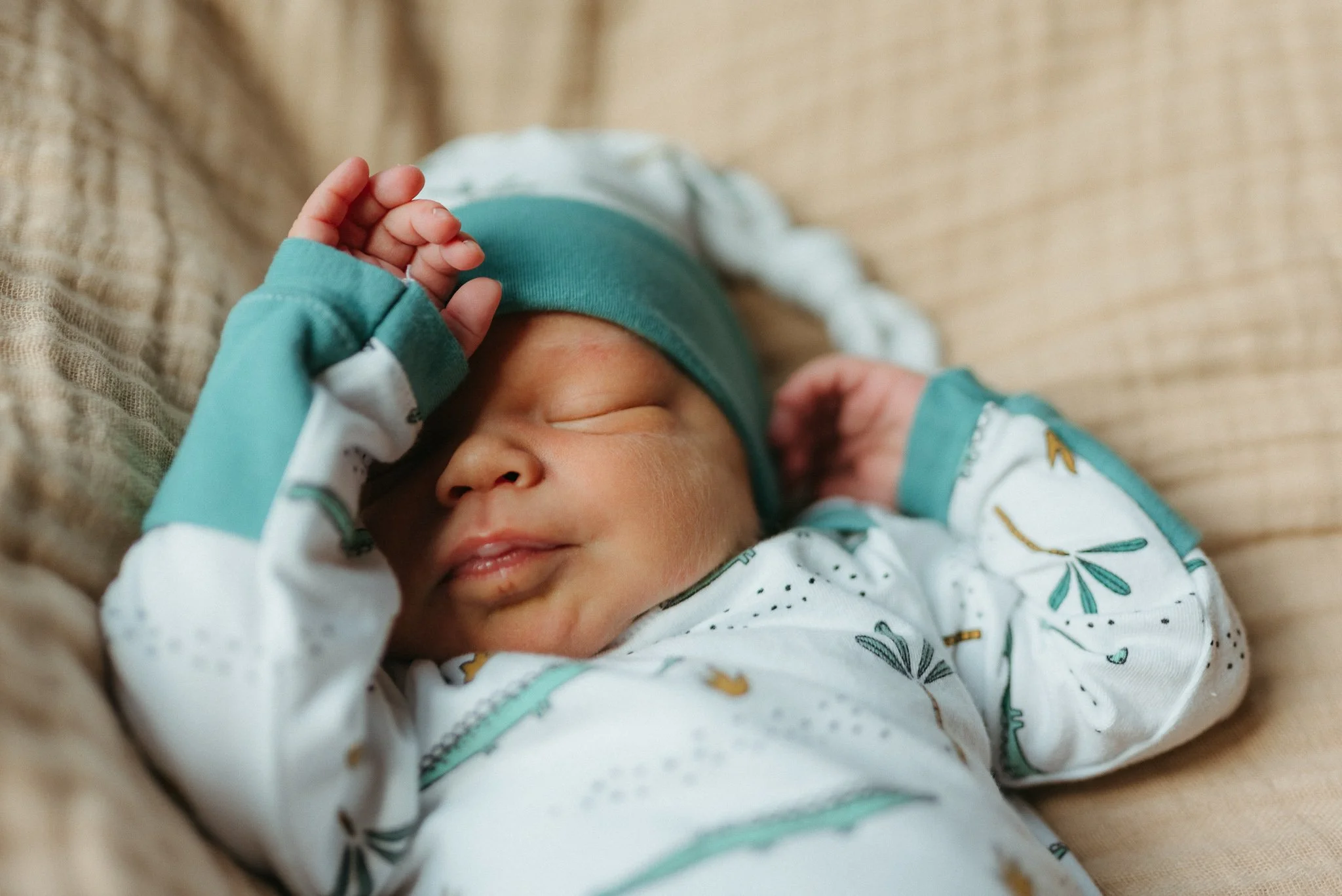 Close-up of a sleeping baby wearing a white onesie with a leaf pattern and a teal hat, with hand resting on forehead.