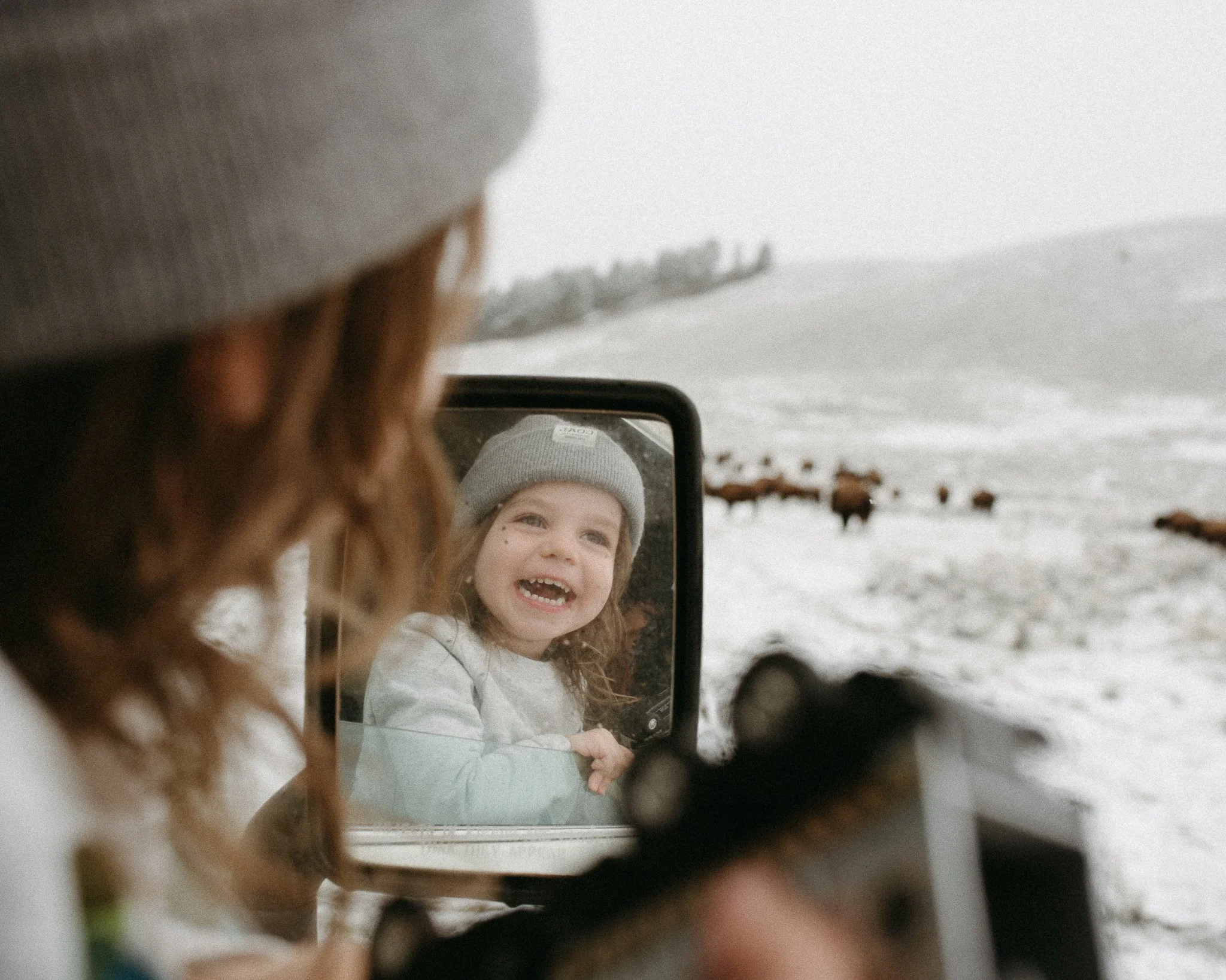 A young girl with curly hair and a gray beanie smiling while looking into a mirror outside in snowy weather, with reindeer in the background.