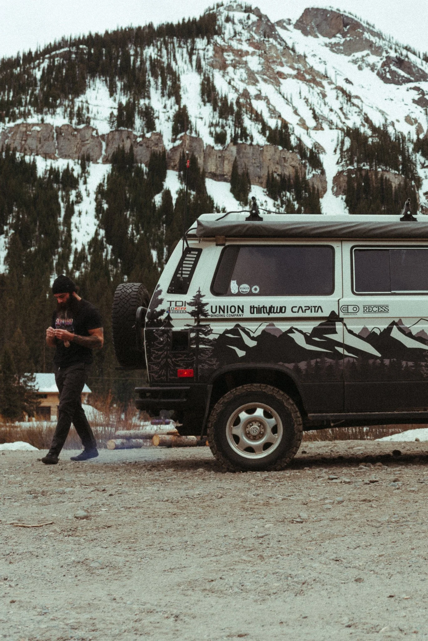 A man in a black shirt and black pants is walking outdoors on a dirt surface, with a large black and white van behind him. The van has mountain and forest decals, and various stickers on the side. In the background, there are tall, snow-covered mount