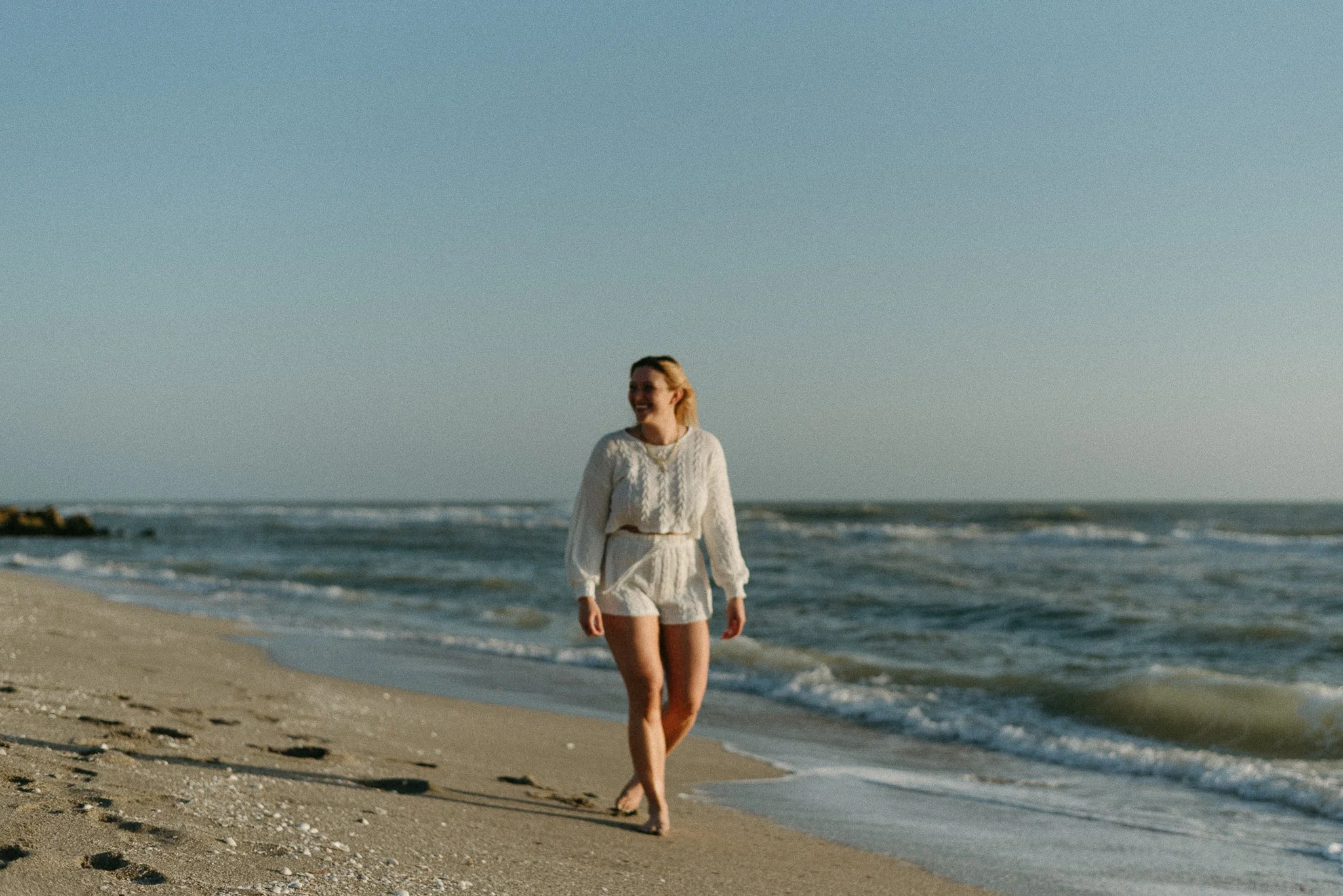 A woman walking on the beach wearing a white sweater and shorts, with the ocean and clear sky in the background.