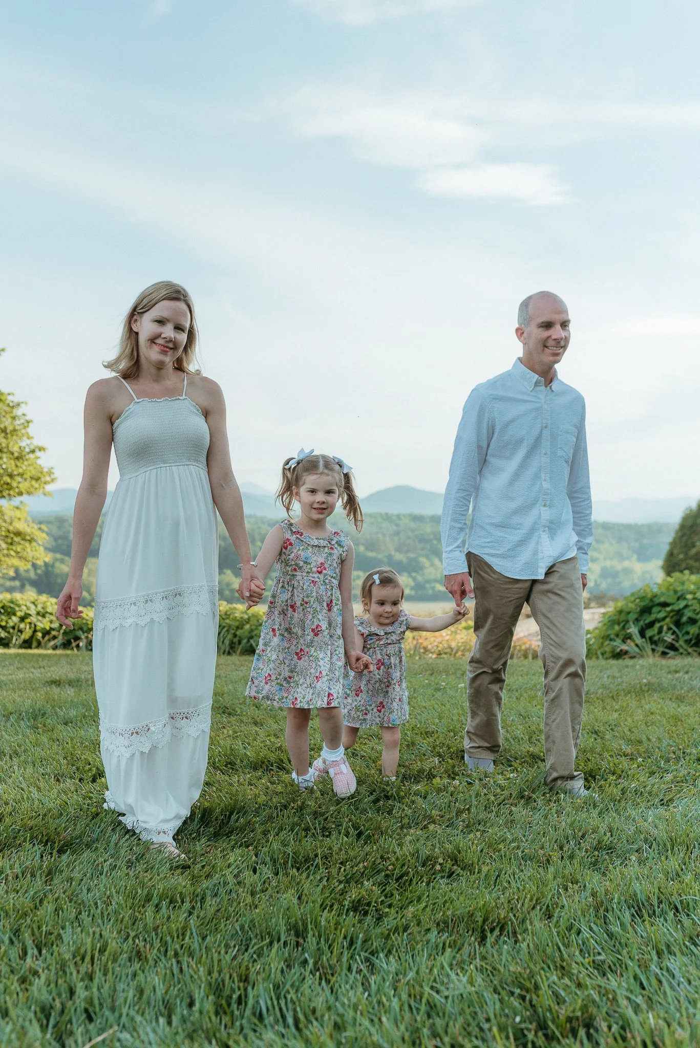 A family of six walking outdoors in a green field with hills and trees in the background. The mother and father hold hands with their three young daughters, all dressed in casual summer clothing.