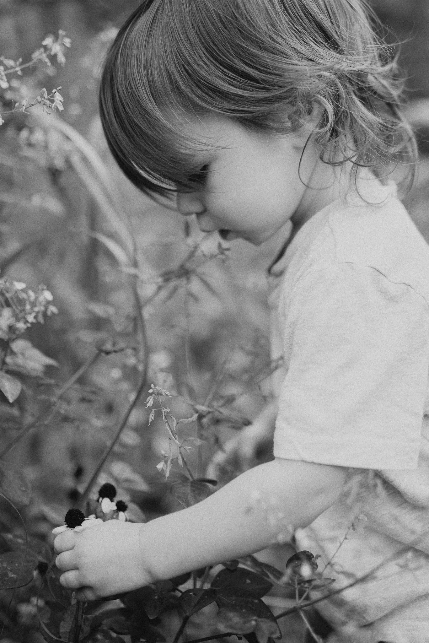 A young girl with short hair in a side profile, cradling a bunch of flowers or plants with both arms, surrounded by leaves and small flowers, in black and white.