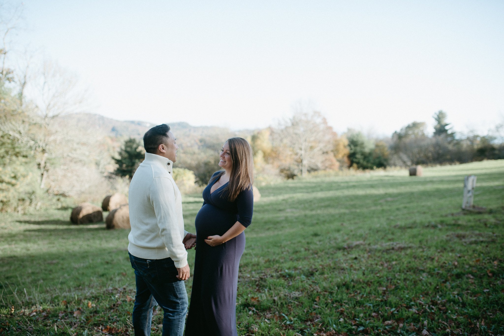 A couple, a man and a pregnant woman, holding hands and smiling at each other outdoors in a park with trees and a grassy field.