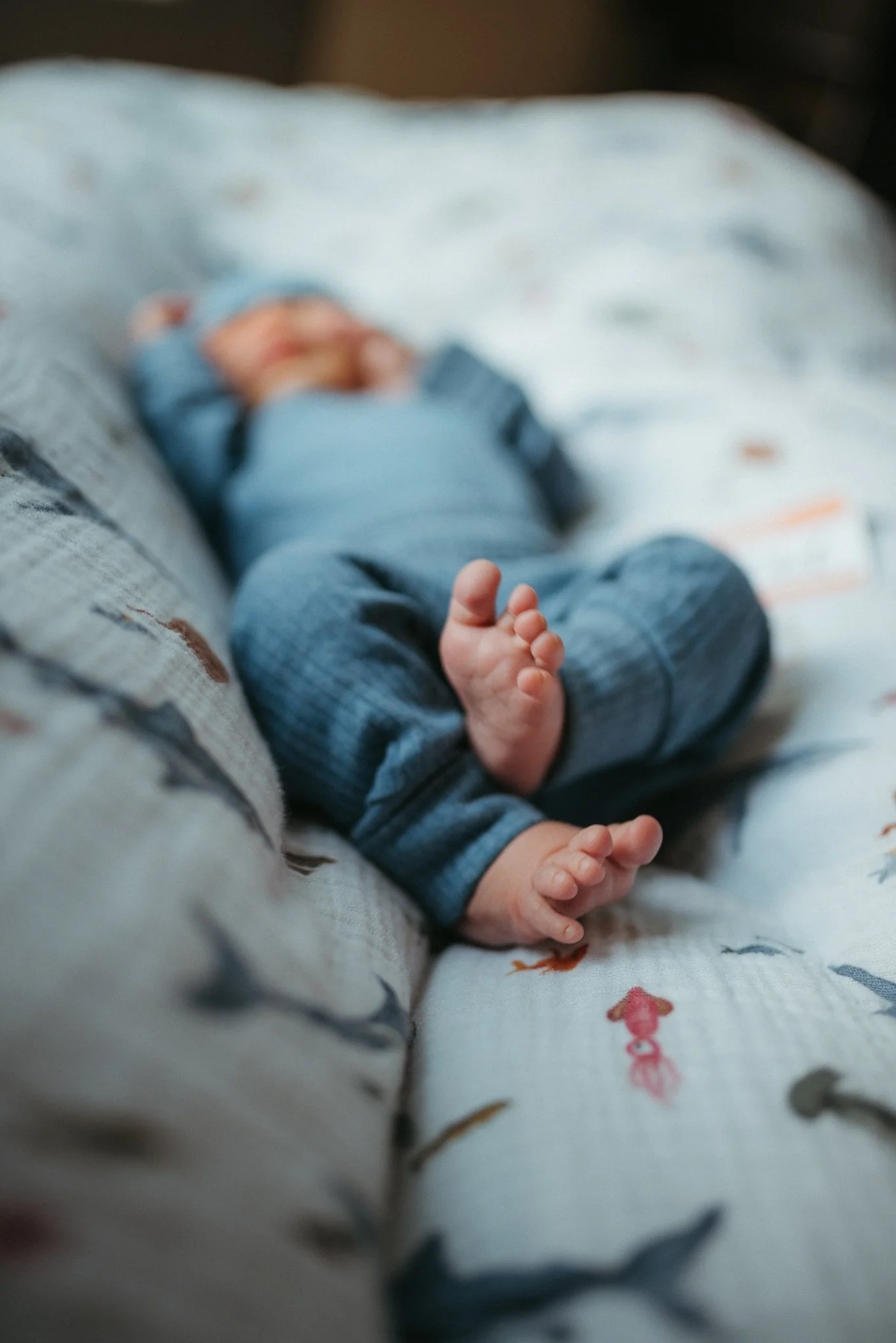 Close-up photo of a newborn baby lying on a bed, wearing a blue onesie, with a soft-focus background of bedding with colorful animal prints.