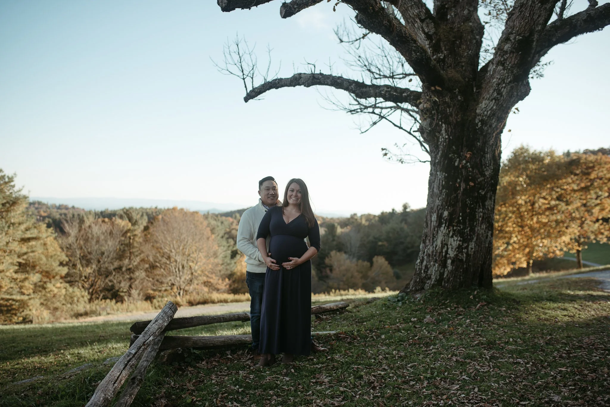 A pregnant woman and a man stand together outdoors in a park during autumn, smiling at the camera. The woman is wearing a long dark dress, and the man is wearing a light-colored sweater. They are near a large, leafless tree, with a landscape of trees