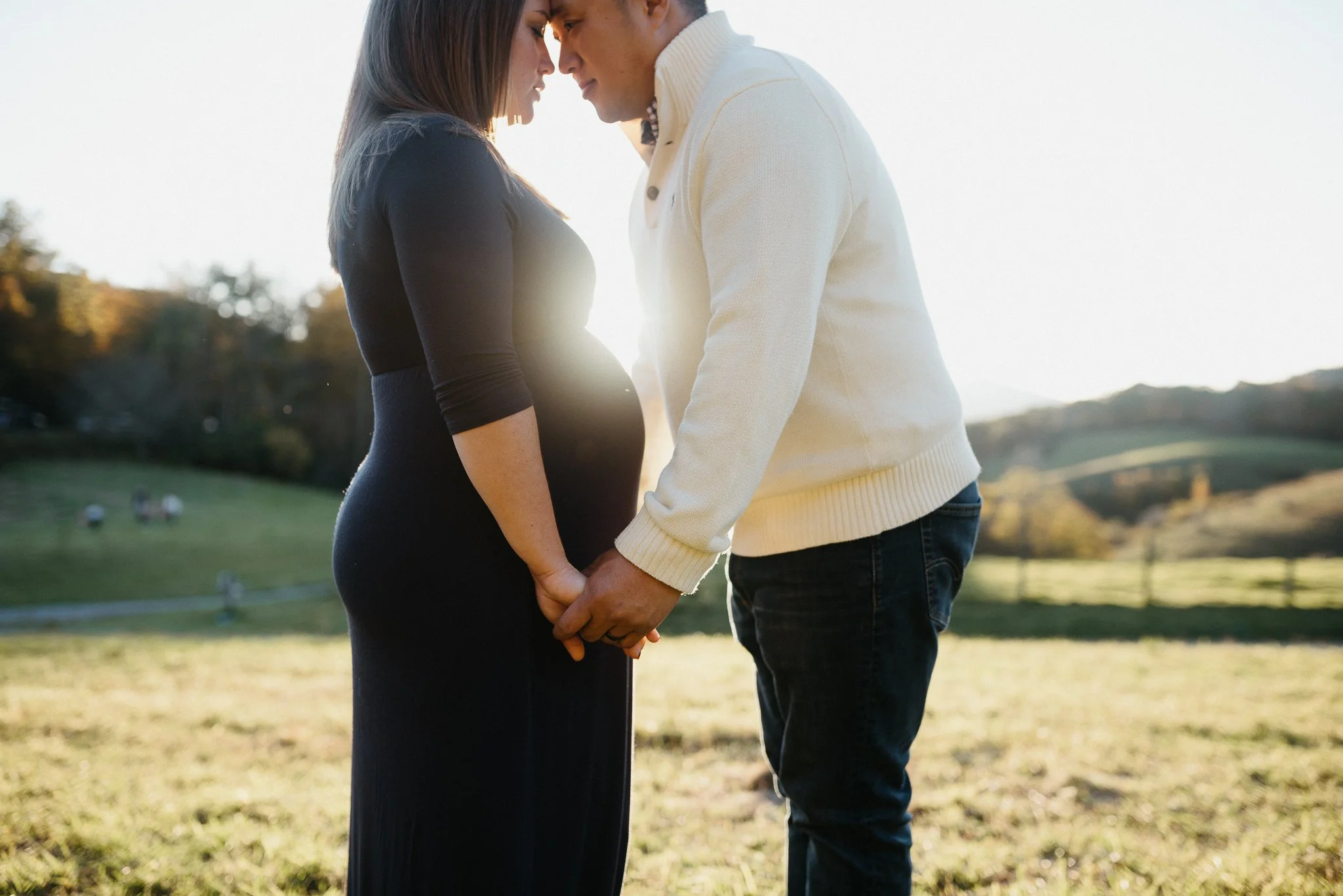 A pregnant woman and a man are standing close together outdoors, touching foreheads and holding hands, with the woman's pregnant belly visible. They are dressed casually, with the woman in a black dress and the man in a white sweater. The scene is ba