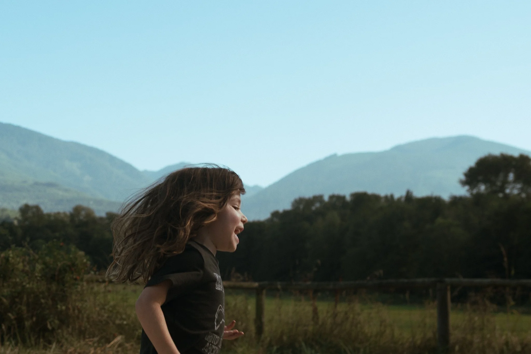 A young girl with long brown hair running outdoors with mountains in the background and a clear sky.