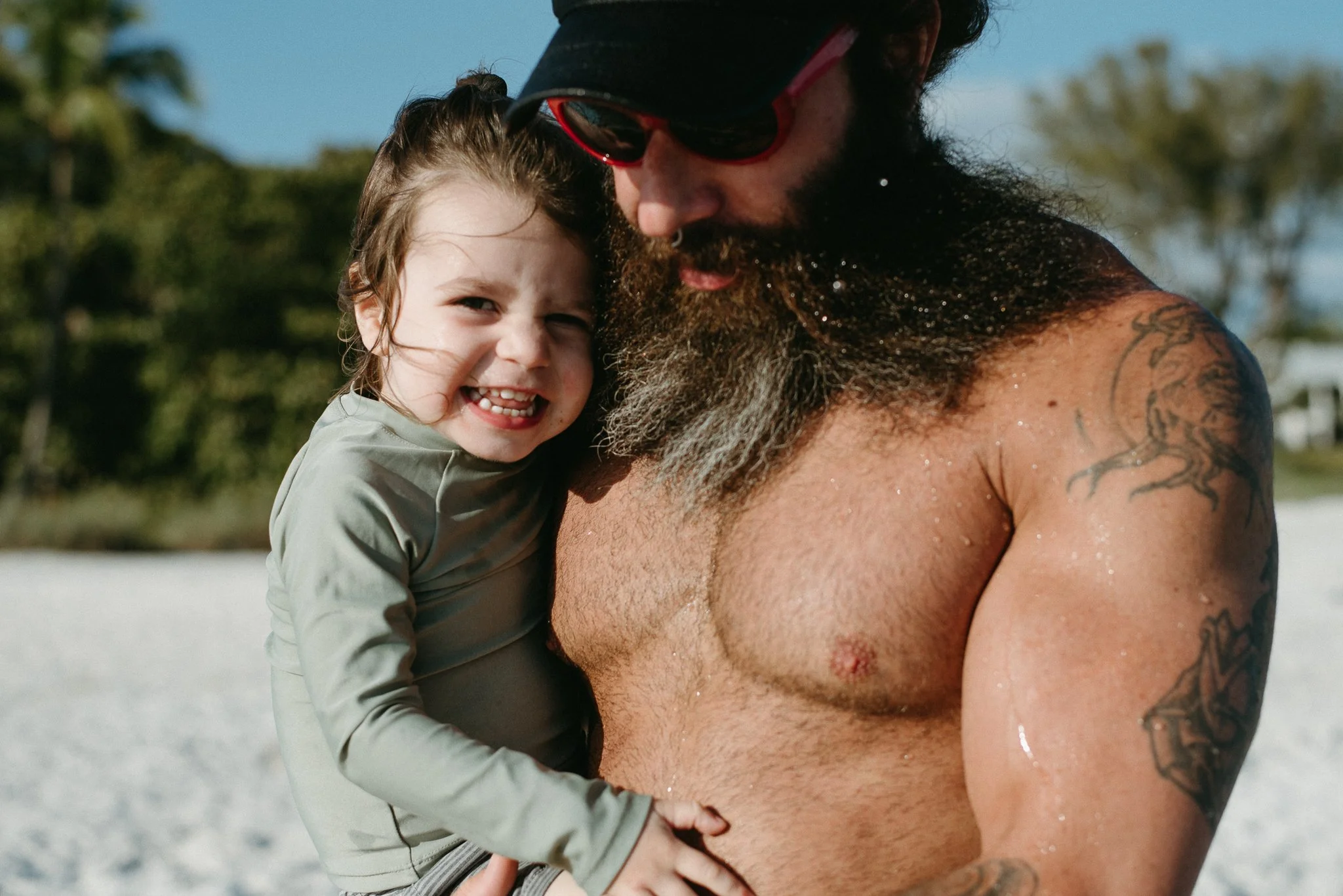A smiling young girl with wet hair hugging a bearded man with sunglasses and tattoos on his arm, both outdoors on a sunny day.