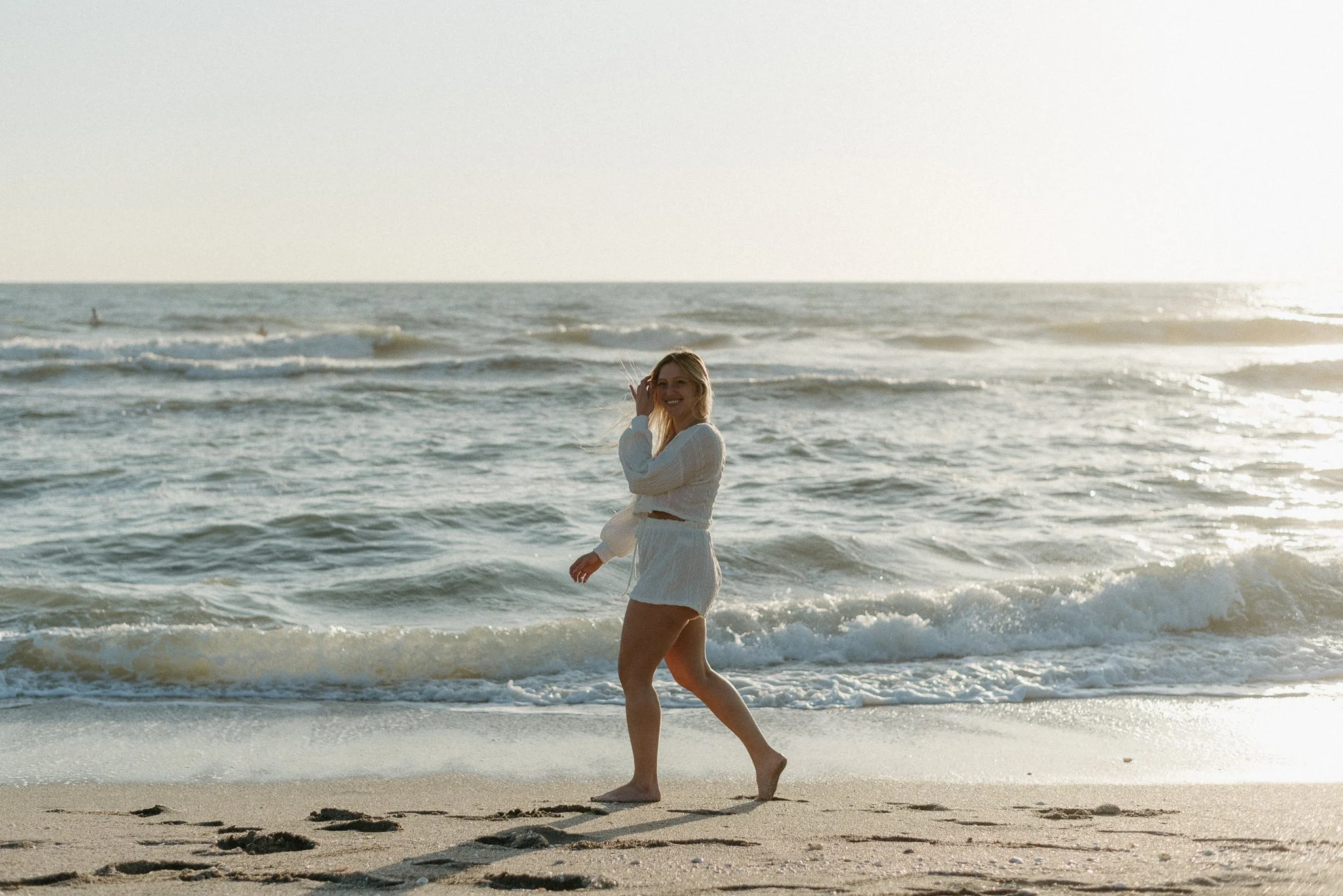 A smiling woman in a white sweater and shorts walking along the shoreline at the beach, with ocean waves and a clear sky in the background.