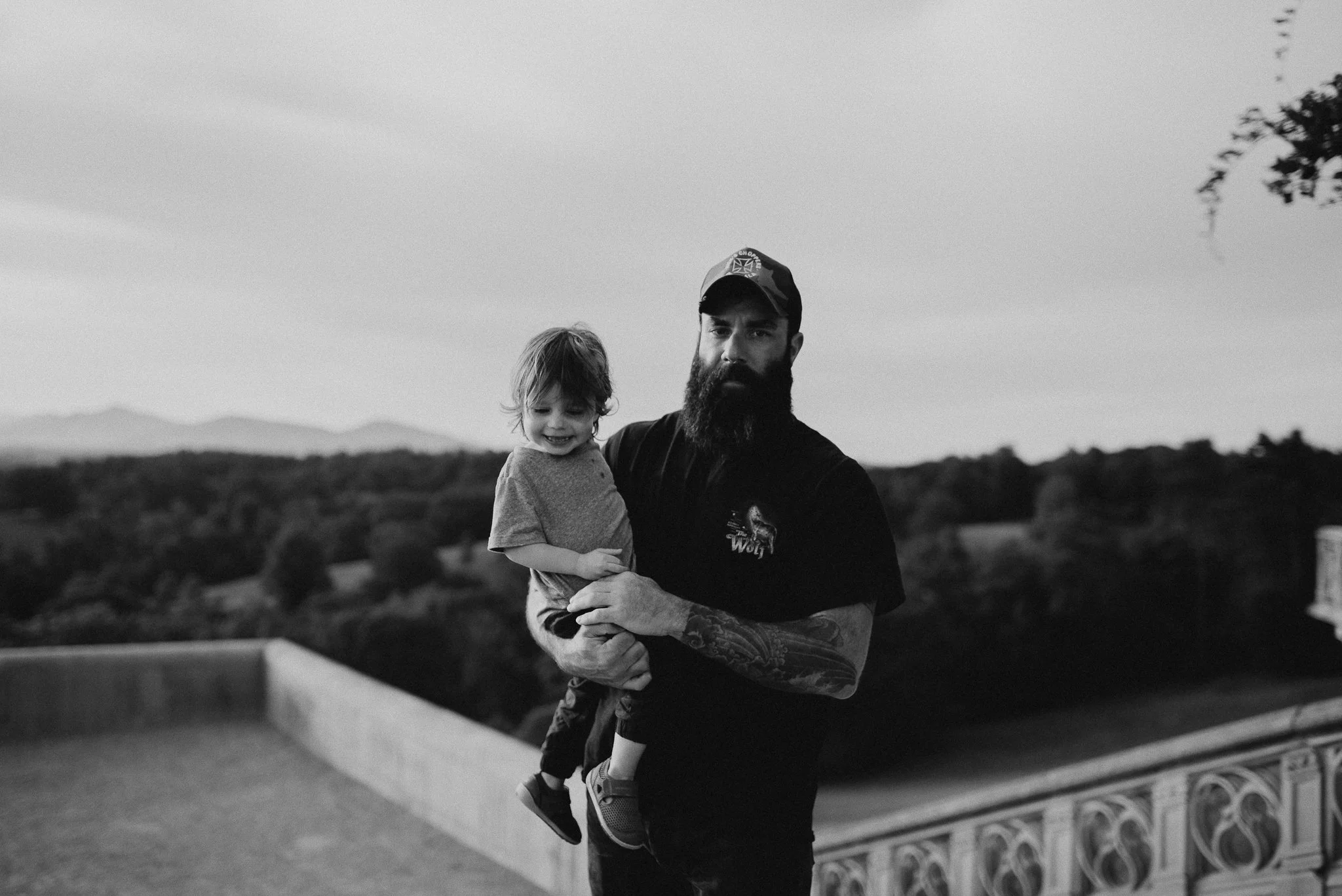 A man with a beard, wearing a cap and a black shirt, holding a smiling girl with curly hair in his arms outdoors during the daytime.