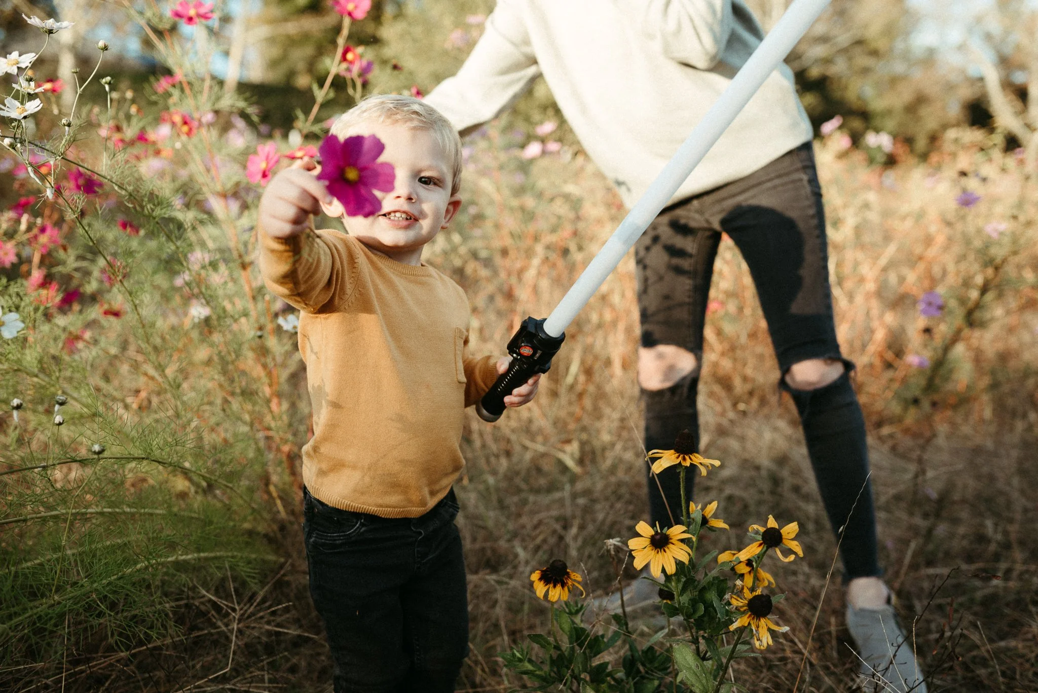 Young boy with blond hair holding a pink flower in a garden with colorful flowers and an adult partially visible in the background.
