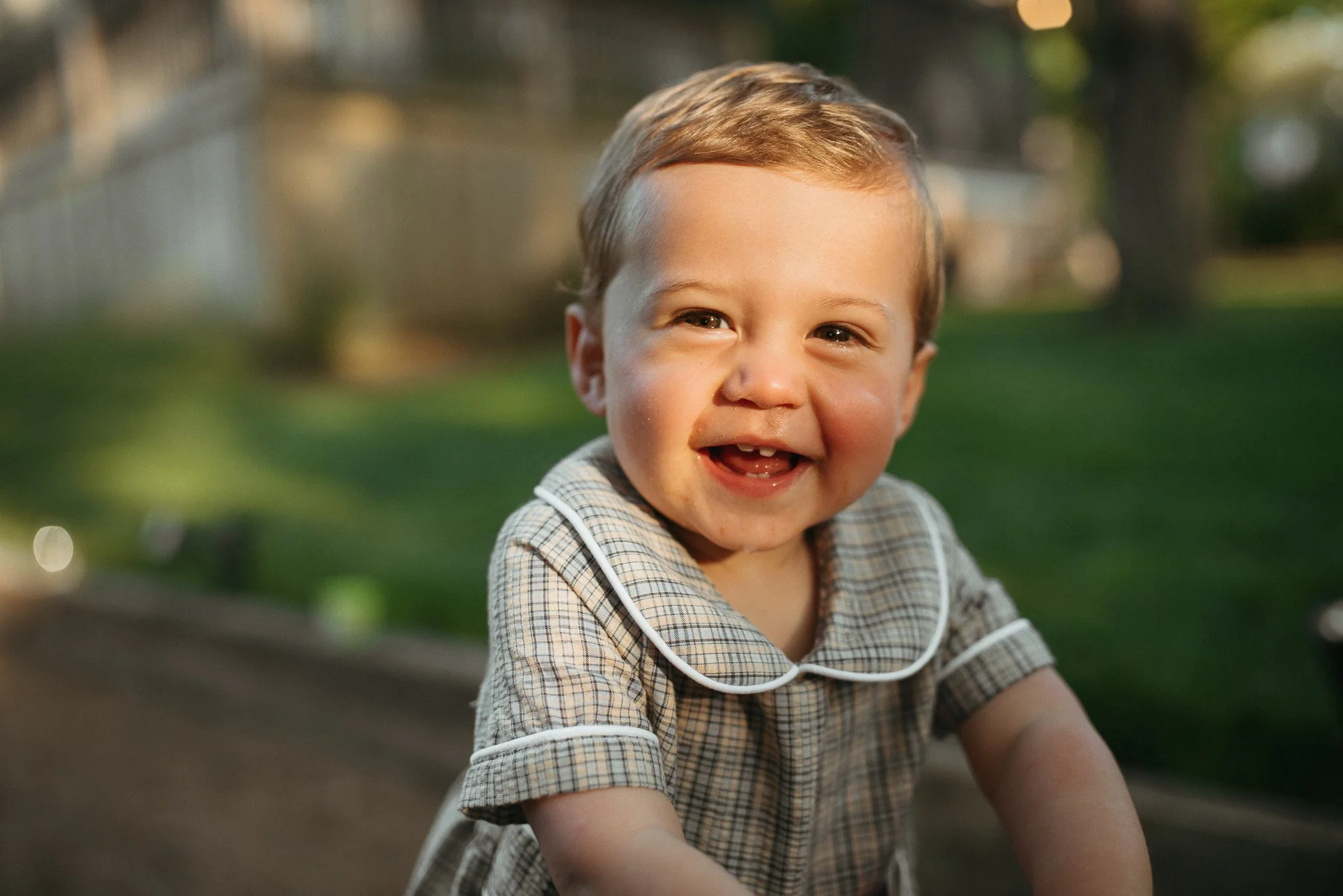 A young boy with light brown hair and a plaid shirt smiling outdoors in a park or garden area.