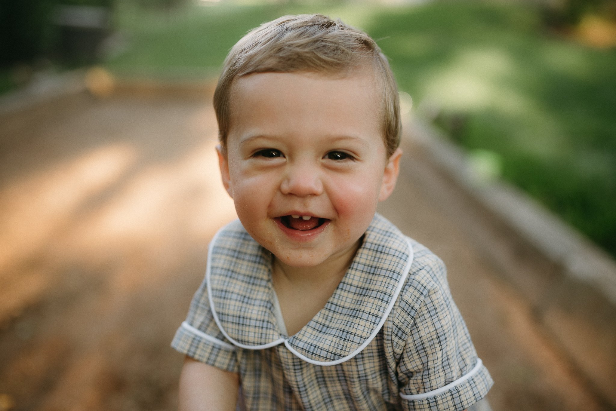 A young boy with light brown hair and a checkered shirt smiling and crouching outdoors on a sunny day with a blurred green background.