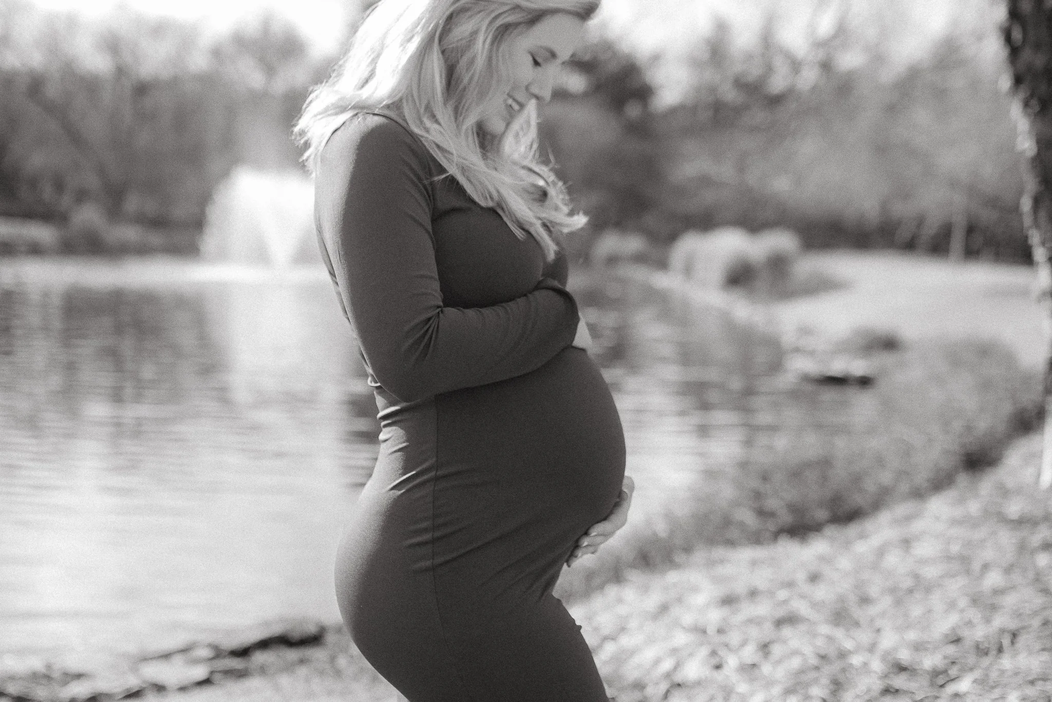 A pregnant woman smiling outdoors near a body of water, with trees in the background, in black and white.